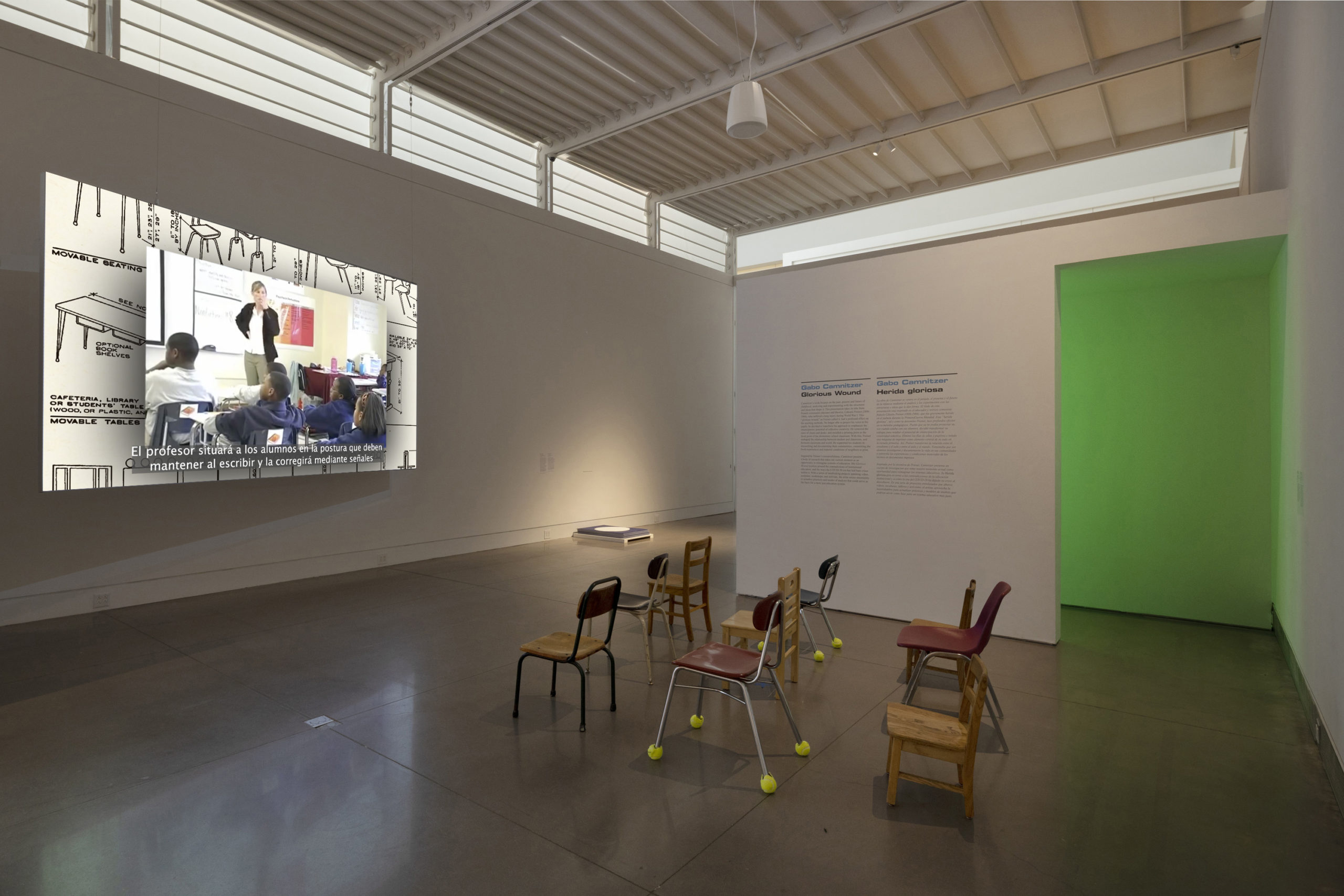 On the left side, there is a large projected still from Gabo Camnitzer’s film, a Picture-in-Picture view of archival classroom materials and blueprints. In the background to the right of the projection, a stack of blue and white posters are on the floor. To the right of the posters, facing the projection, are nine small public school chairs in different colors and materials. Behind the chairs is a large wall text of the exhibit, and to the right is an empty doorway emanating bright green light.