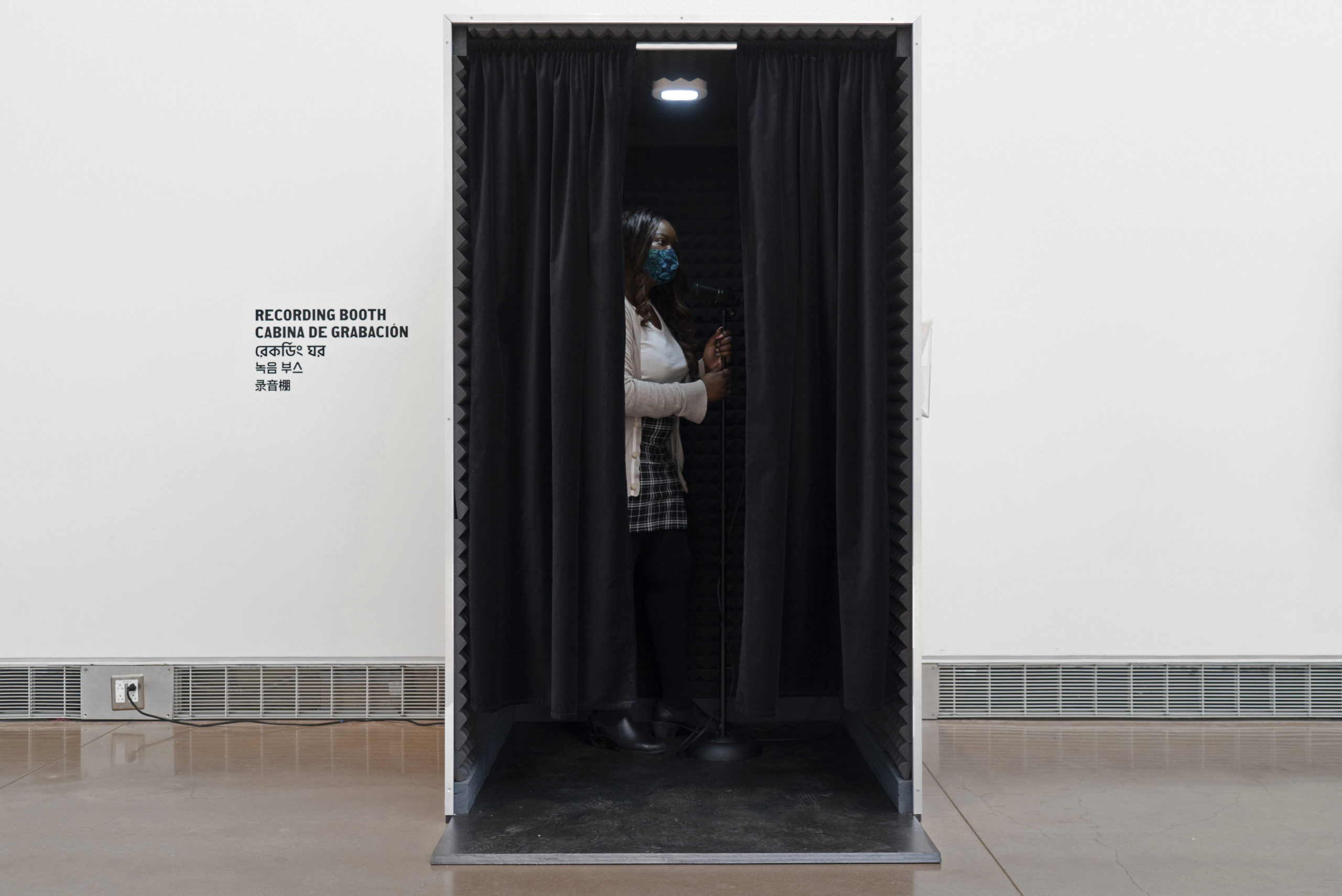 In the center, a visitor is standing in a wooden rectangular recording booth with a black curtain split open. On the wall, to the left of the recording booth reads Recording Booth.