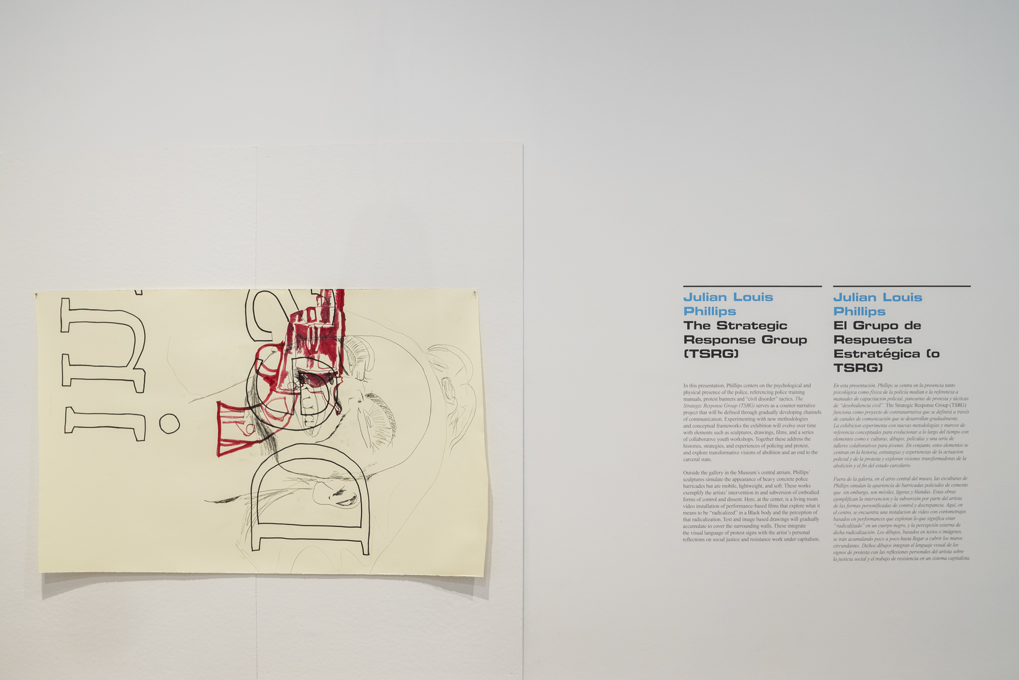 On the left, a drawing by Julian Louis Philips in mixed mediums is pinned to the wall horizontally. On the right, there is the gallery wall text reading the artist’s name Julian Louis Philips, the exhibition title The Strategic Response Group, and information describing the exhibition and the artist’s work.