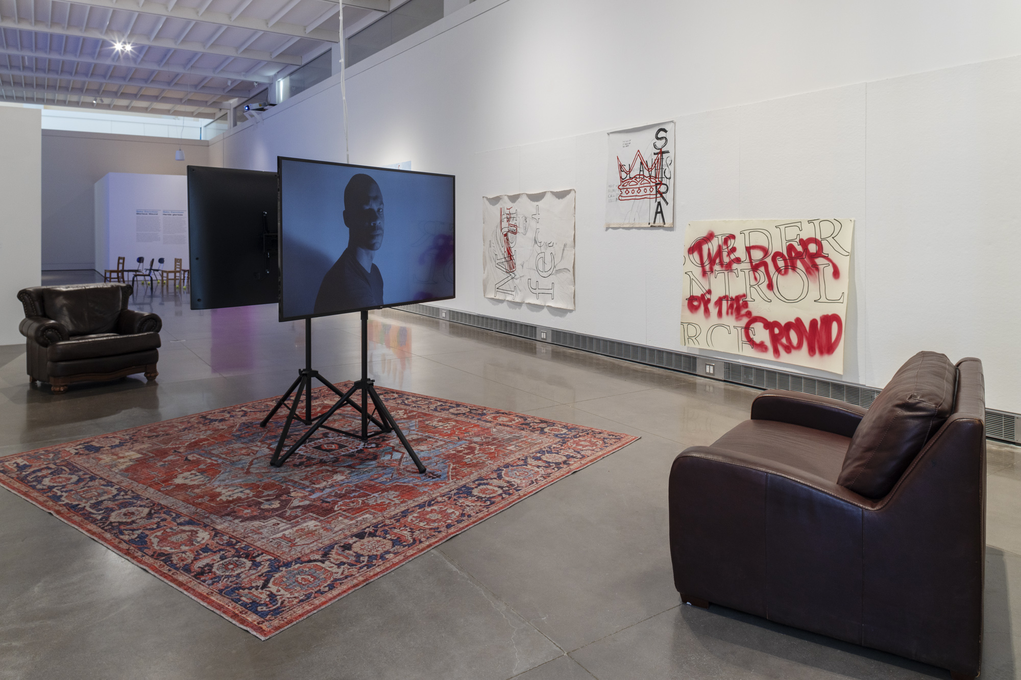 An exhibition space with text and image based art hanging on the back wall. In the center is two, flat screens on tripods facing back-to-back. The screens are centered on a red, oriental rug, in between two, brown leather armchairs. On the screen facing forward is a man against a white backdrop. The shot of him is from the bust up and he is glancing over shoulder with a still facial expression.