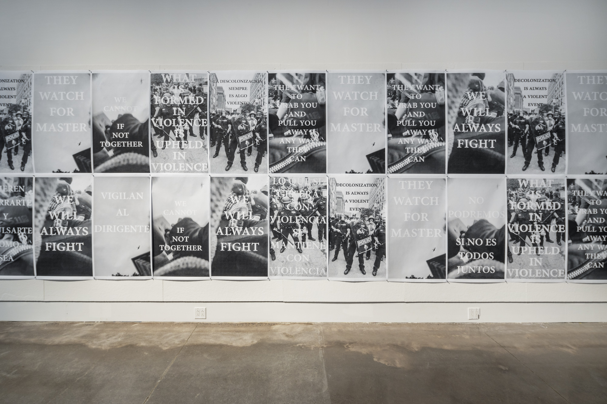 Two rows of black and white postered installed on a white exhibition walls. The posters are made up of black and white photographs of police barricades formed at protests, a city sky, and protestors. Over the photographs are a series of phrases including “We Will Always Fight”, “Decolonization Is Always A Violent Event”, “What Is Formed In Violence Is Upheld In Violence”, “They Watch For Master”, and “We Cannot If Not Together” in both English & Spanish.
