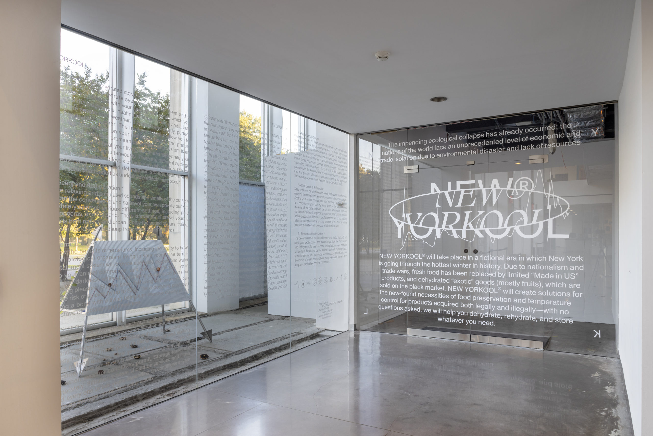 A view of two clear walls in an exhibition space that have with white, vinyl text. The longer wall on the left has three columns of paragraphs stickered on. The wall adjacent, to the right, has the exhibition title “New Yorkool” distorted and arranged along an oval. Above and below the centered exhibition title is a show statement.