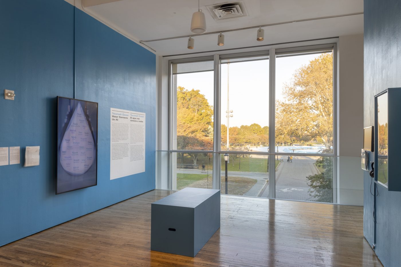 A bright room has two blue walls on opposite sides, and a large floor-to-ceiling window wall in between looking out on the green trees and gray pathways of Flushing Meadows Corona Park. A simple box/bench in between is painted the same blue as the walls. The left wall has a large monitor displaying a water droplet with text inside, and to the right of the monitor is a large wall text. The right wall has a glass vitrine, a small monitor, and smaller descriptive text.