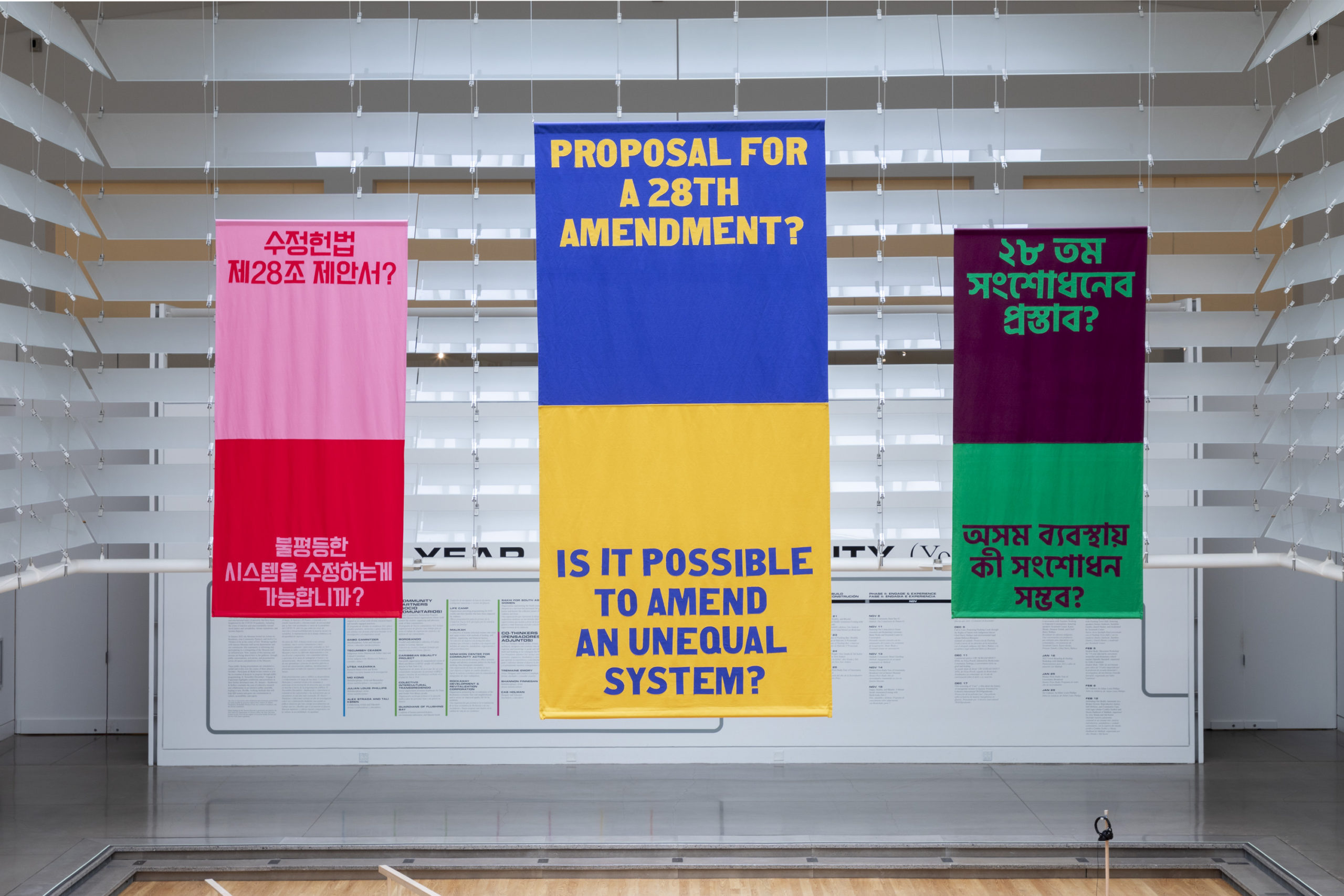 Three colorful banners hang from the ceiling reading Proposal for a 28th Amendment? And Is it possible to amend an unequal system? In Korean, English, and Bengali.