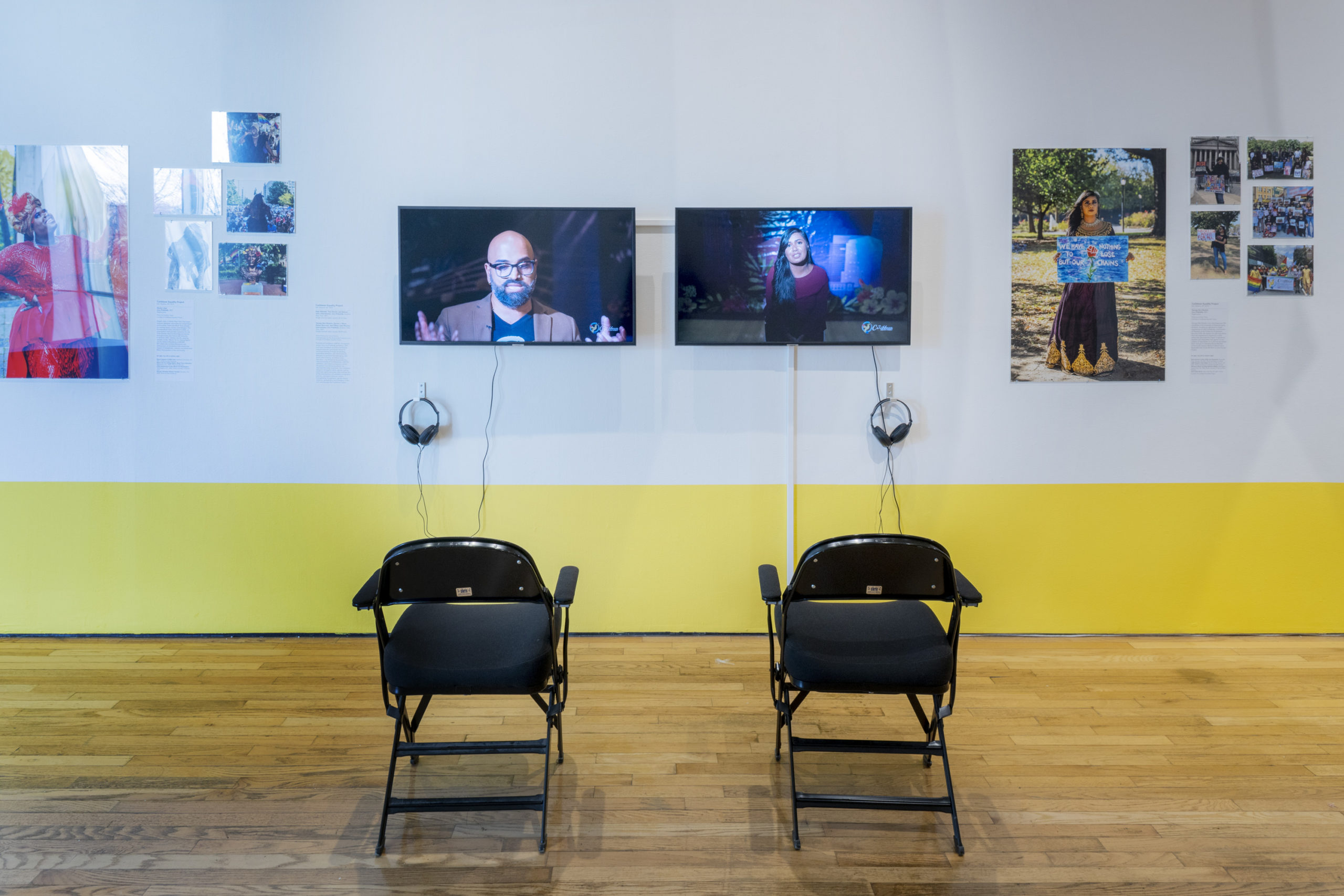 Two black chairs placed in front of an exhibition wall. The exhibition wall is white with a yellow strip at the bottom. In the center, two screens with headphones have been installed. The screens are displaying two speakers. Framing the monitors is community member portraits and photographs of community events from The Caribbean Equality Project.
