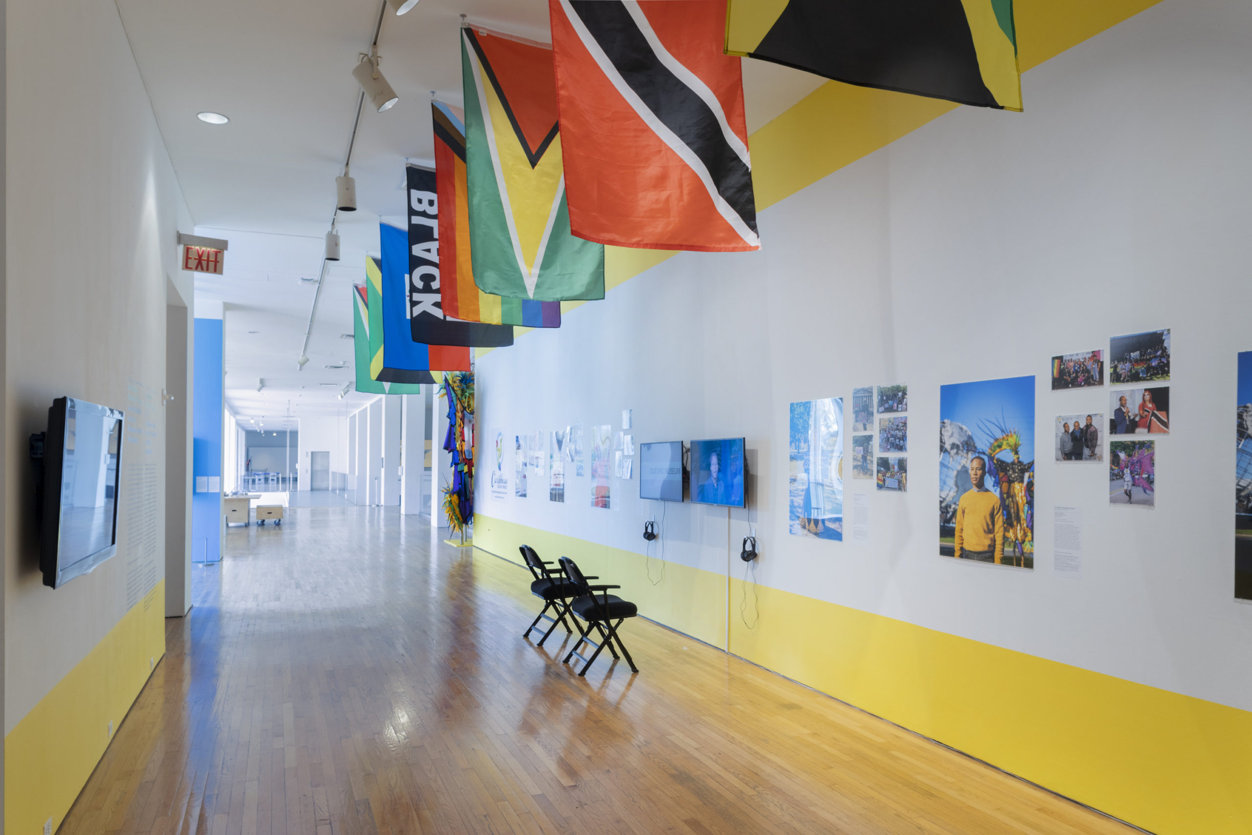 A exhibition hallway with white walls bordered with a yellow stripe at the top and bottom. The wall on the left has exhibition text and a monitor. The wall on the right has two monitors, portraits of community members and photographs of community events from The Caribbean Equality Project.