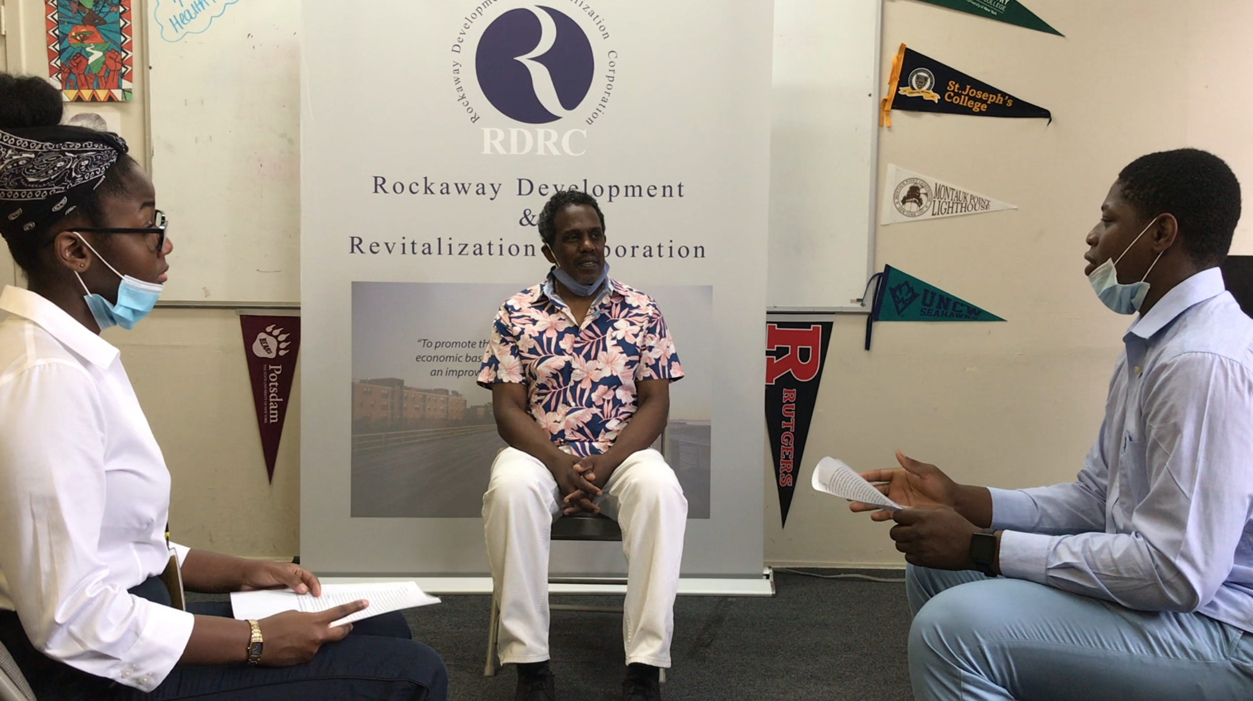 Three Black adults sit in chairs forming a circle in what appears to be a classroom. They are dressed in business causal wear and have surgical masks on but tucked under their chins for a conversation. Behind them is a large poster with the logo for the Rockaway Development & Revitalization Cooperation.