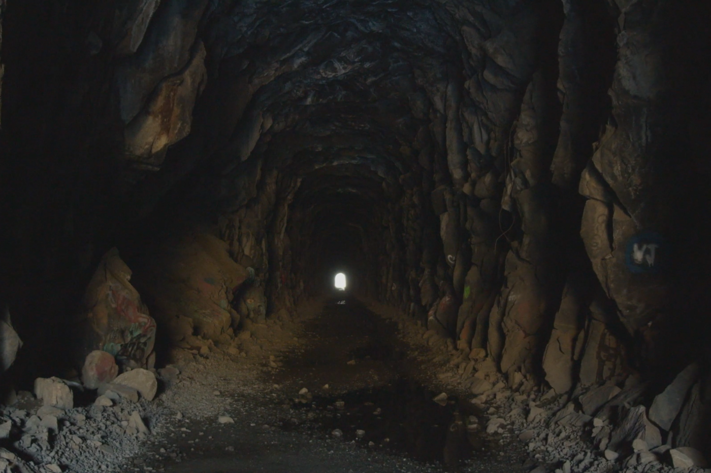 A long narrow tunnel within a rock formation. At the end of the tunnel is an opening shaped like a beam of light.