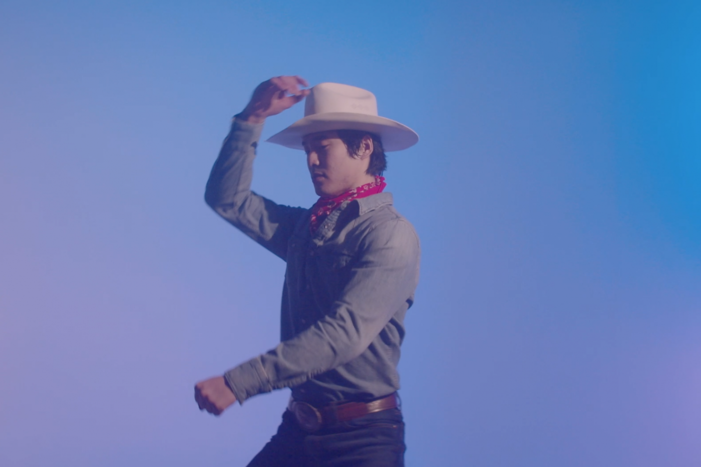 An Asian man dressed in blue jeans, a jean collared shirt, a brown cowboy belt, a red bandana around his neck, and a white cowboy hat poses centered against a blue background. His arms are gesturing as if he is riding a horse.