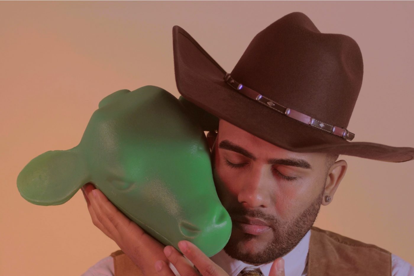 A tight shot of an Asian man dressed in a white collared shirt, brown suede vest, and brown cowboy hat. He has dark brown eyebrows, a close cut bread, and is wearing stud earrings. Perched on his shoulder, gently propped up by his hands, is a green mold of a cow’s head. He is tilting his head gently with his eyes closed so that his head and the cow’s head together create the shape of a heart.