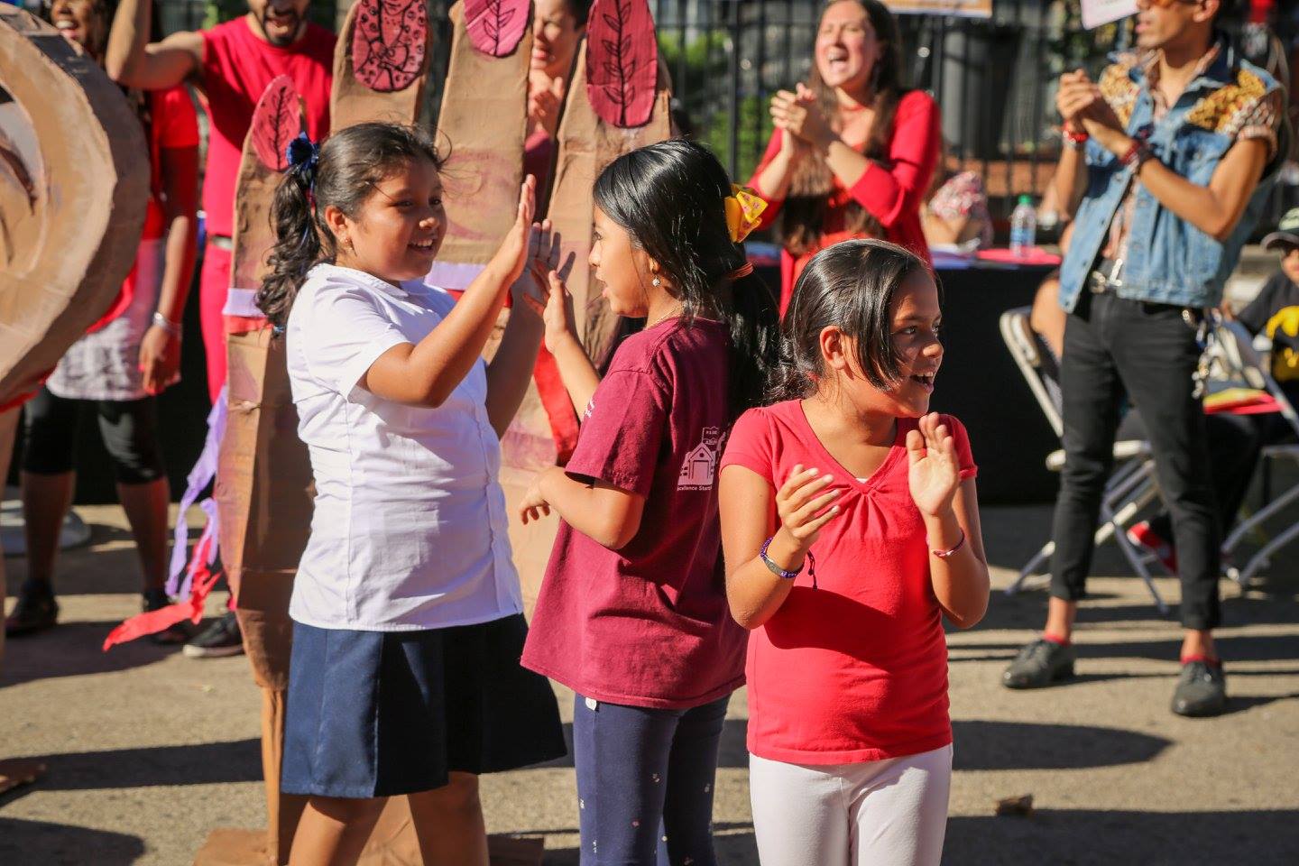 A crowd of inter-generational Latinx community members stand outside for a community arts event. Focused in the foreground is three young girls. Two are playing a hand clapping game and one is cheering. Behind them is a papier-mâché sculpture of a hand and adults also cheering and clapping.