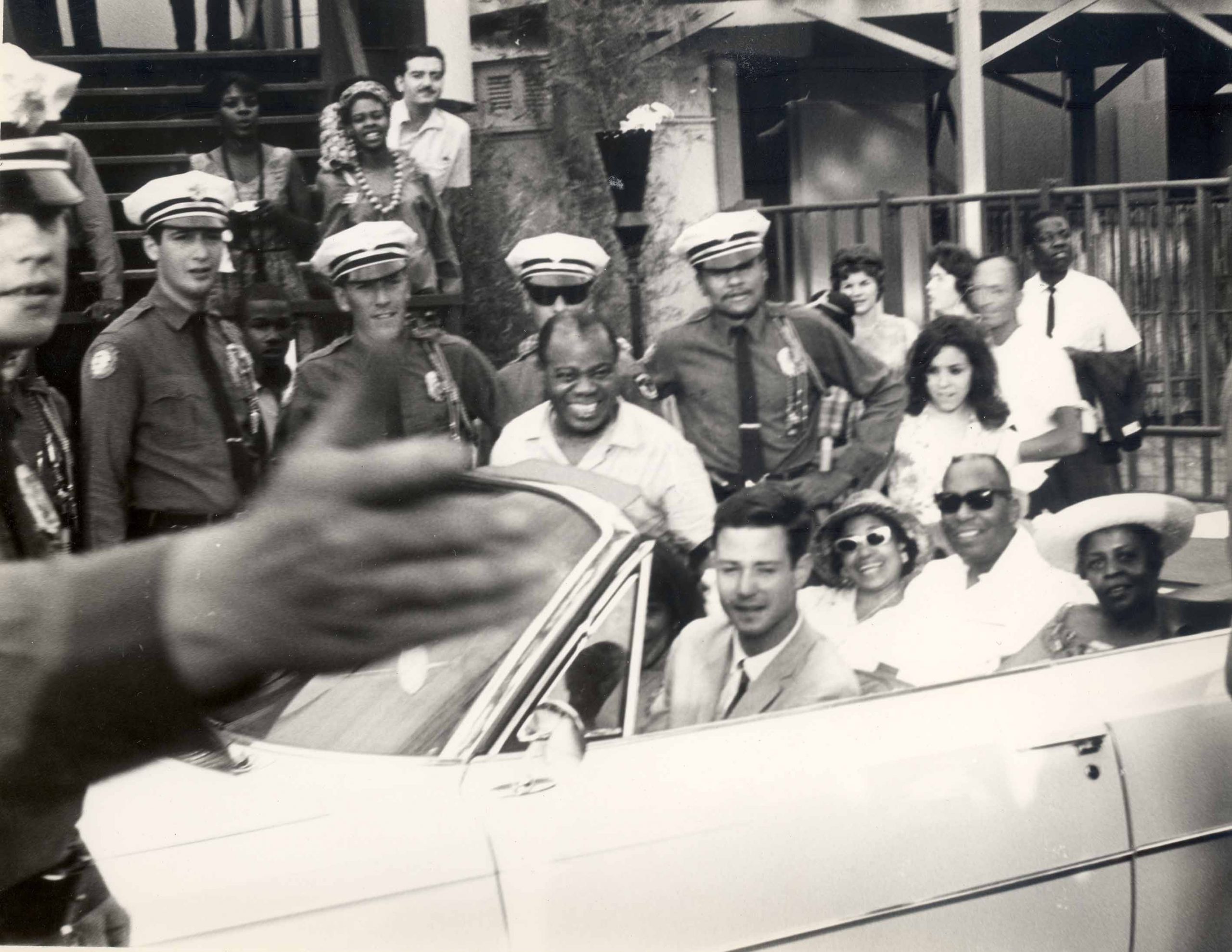 A black and white photograph of Louis Armstrong cheesin’ his pearly whites. His gaze is towards another camera lens. Behind him is a row of police officers. Together, with a crowd of onlookers, they are captured standing. While the police officers are on the curb, Louie is crouching up out of a parked convertible, filled with seated passengers.