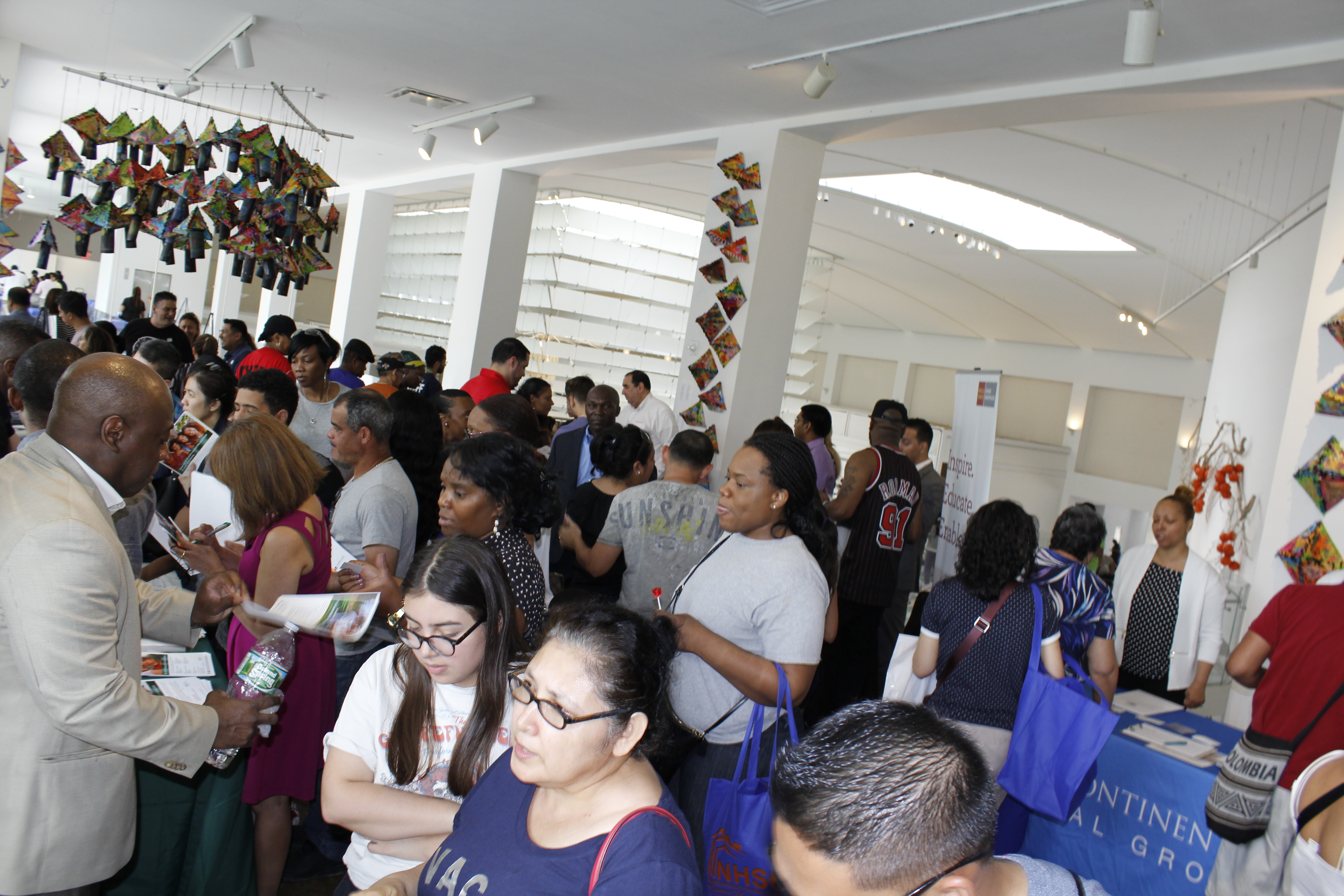 A large crowd of people inside Queens Museum interact with information tables at the Neighborhood Housing Services of Queens CDC 2018 Housing Fair.