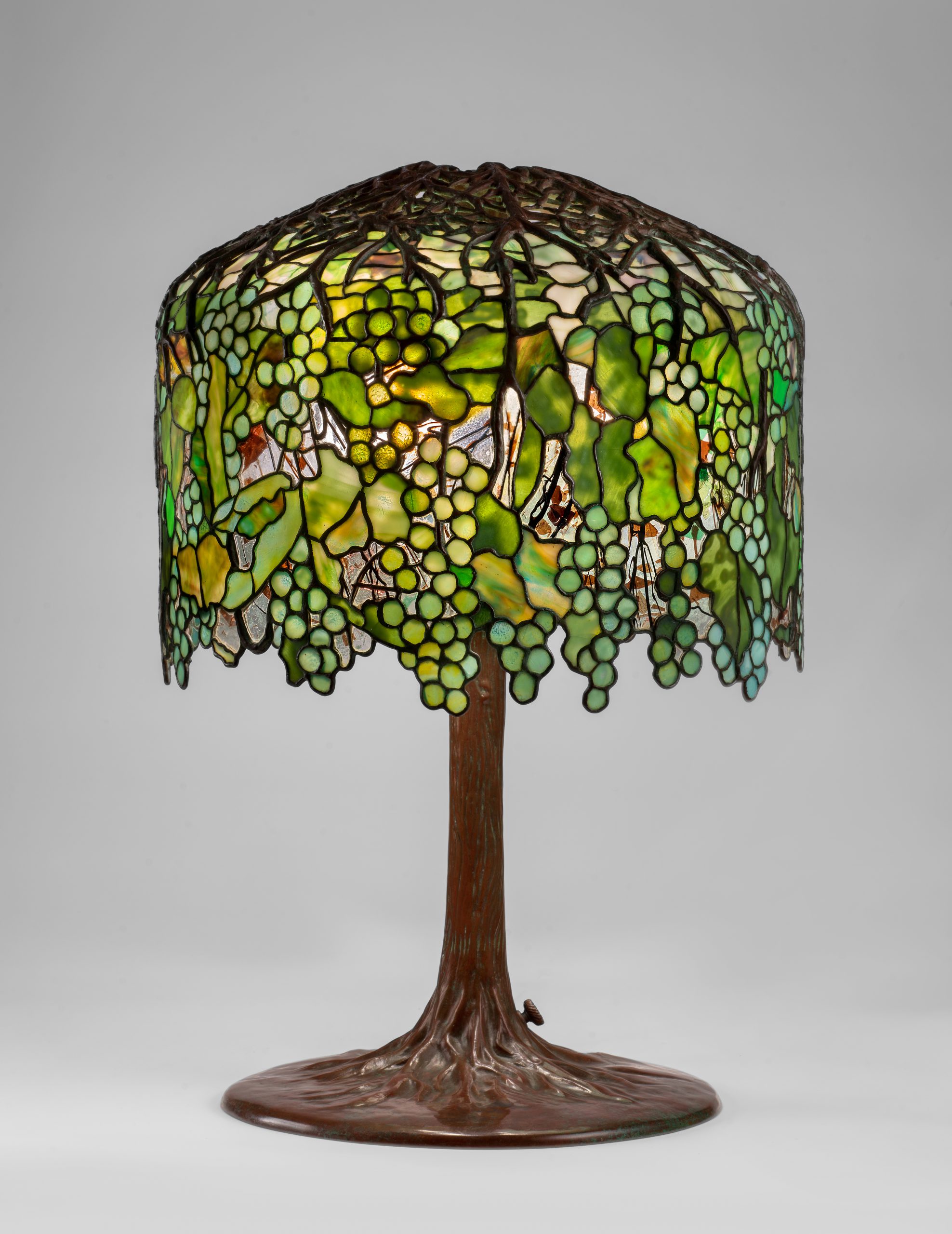 A lit lamp with a brown base that resembles a tree trunk. The base extends into the lampshade as brown branches. The lampshade is made of organic shaped mosaic pieces, in varying shades of green and yellow.