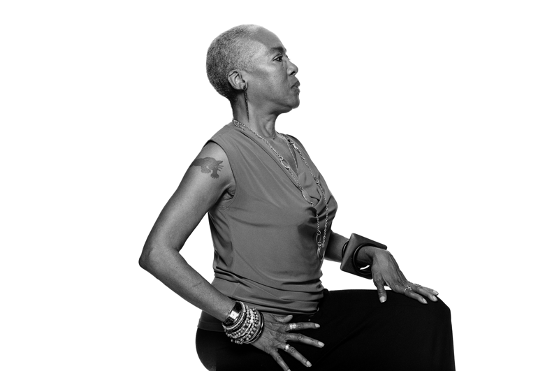 A side profile portrait of a Black woman in black and white. She is posing against a white backdrop and in a seated posture. One hand is on her hip and the other is on her thigh, both clearly displaying a collection of bangles and rings. She is wearing black pants, a sleeveless top, a chain necklace and dangle earrings. Her hair is worn in a close cut shave. On her right bicep is a black panther tattoo.
