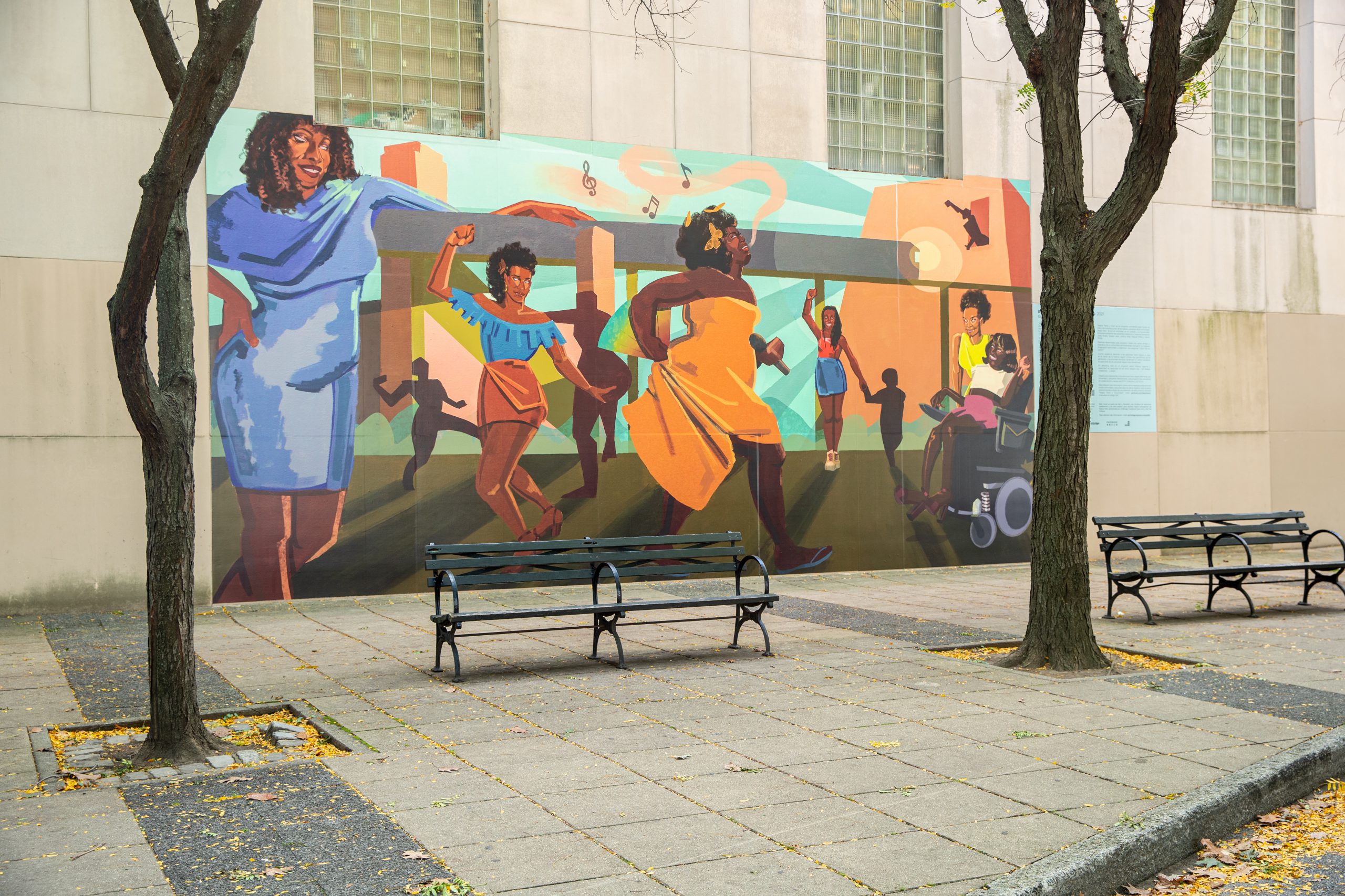 A colorful mural on the sidewall of the museum portrays six Black Trans Femme icons. Marsha P.Johnson, Miss Major Griffin Gracy, Cayenne Doroshow, Qween Jean, Tourmaline, and Gia Love. The first person prominently stands out on the left side in a vibrant blue dress draped across the shoulder, while the person next to them wears a brown wrap skirt and ruffle blue top with one arm up in a fist. In the center is a person in a strapless bright orange dress holding it at the corner as they walk with an orange butterfly in their afro textured hair and music notes leave their lips. The next person is wearing a short blue skirt with a red top with a fist also in the air holding hands with a shadow of a smaller person. The last two people are, a person wearing a pink skirt with a white top in a motorized wheelchair and another person standing over their shoulder wearing yellow.