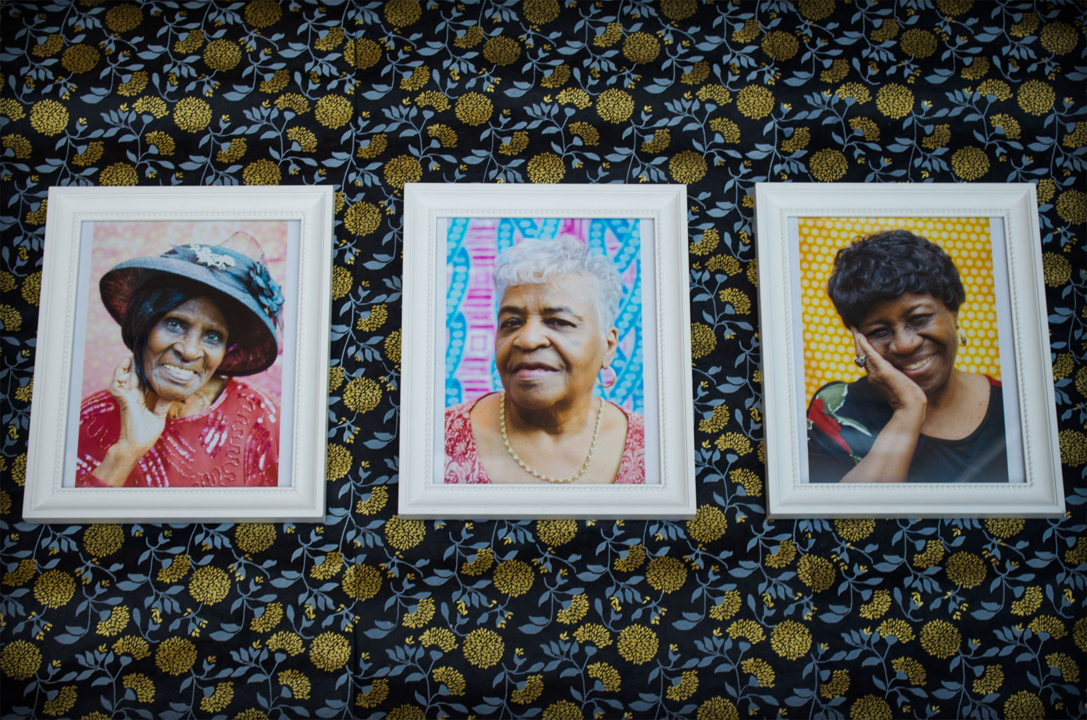 Three framed portraits of Black elderly women. They are each dressed elegantly and smiling against colorful patterned backgrounds. The framed portraits are sitting on a black, blue, and gold floral background.