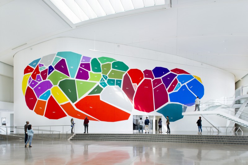 A two story mural of a blob made out of polygons in different, shapes, sizes, and colors. Down at the floor of the exhibition space are adults viewing the mural. Descending from the top right is a twisting flight of stairs.