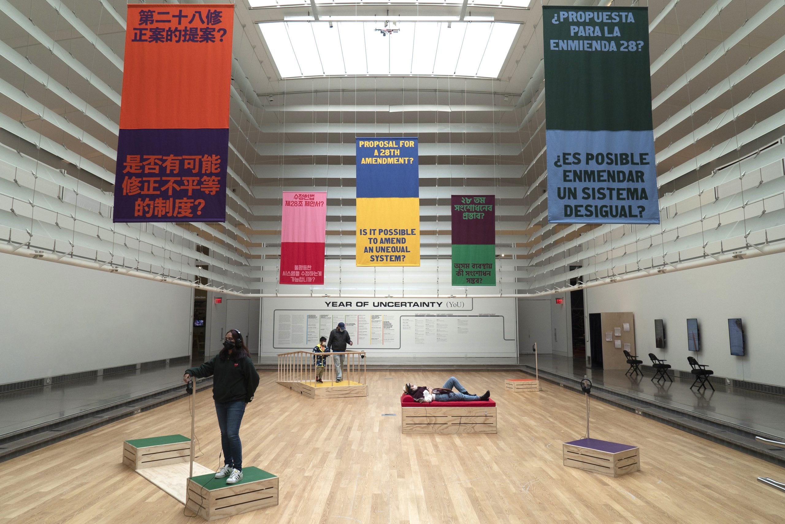 Five colorful banners hang from the ceiling reading Proposal for a 28th Amendment?” And “Is it possible to amend an unequal system? in the five most spoken languages in Corona, NY. Below the banners, four visitors stand and lay on the five wooden colorful soapboxes in different arrangements.
