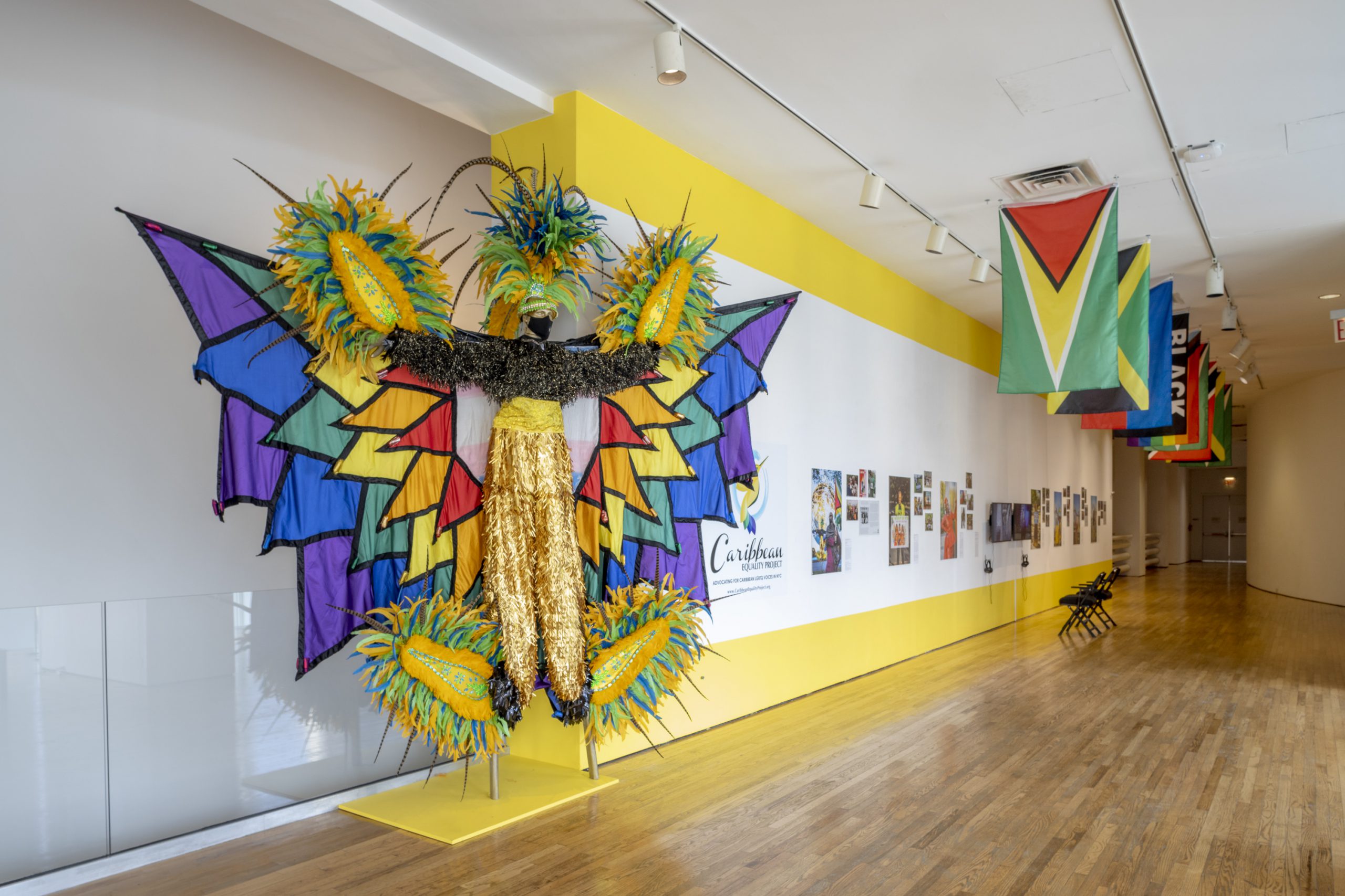 Down a hallway, Photographs, news articles and video installations hang on a yellow and white wall with different Caribbean country flags, a rainbow LGBTQ+ flag, and Black Lives Matter flag, hanging from the ceiling in a row parallel to the wall. At the beginning of the hallway is a mannequin wearing a festive, bright colored, colorful, feathered, carnival outfit.
