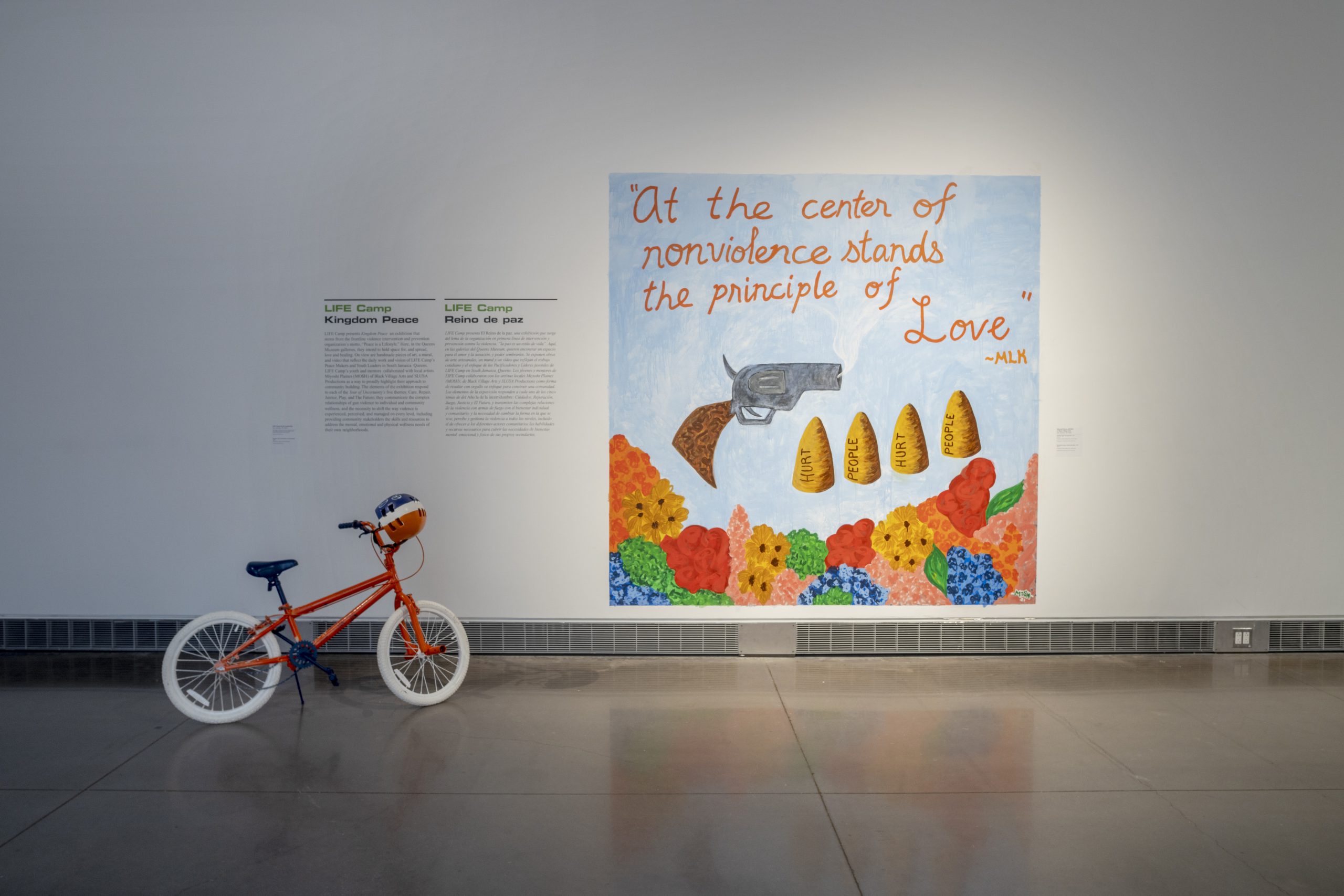 A white exhibition wall with a mural. The mural has a soft blue background and a bed of orange, blue, yellow, and pink flowers at the bottom. Hovering above the flowers is a smoking gun and four large bullets with the phrase “hurt people hurt people” written across them. Above that is a quote by MLK that reads “At the center of nonviolence stands the principle of Love” in orange cursive.