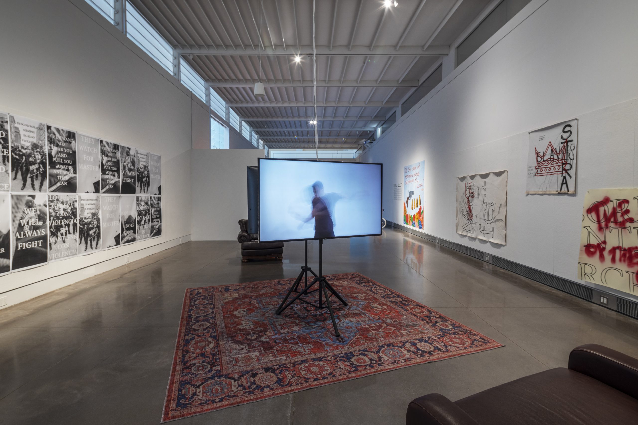 An exhibition space with text and image based art along its two longest walls. In the center is two, flat screens on tripods facing back-to-back. The screens are centered on a red, oriental rug, in between two, brown leather armchairs. On the screen facing forward is a man against a white backdrop. His form is blurred and gestural in a way that implies movement.