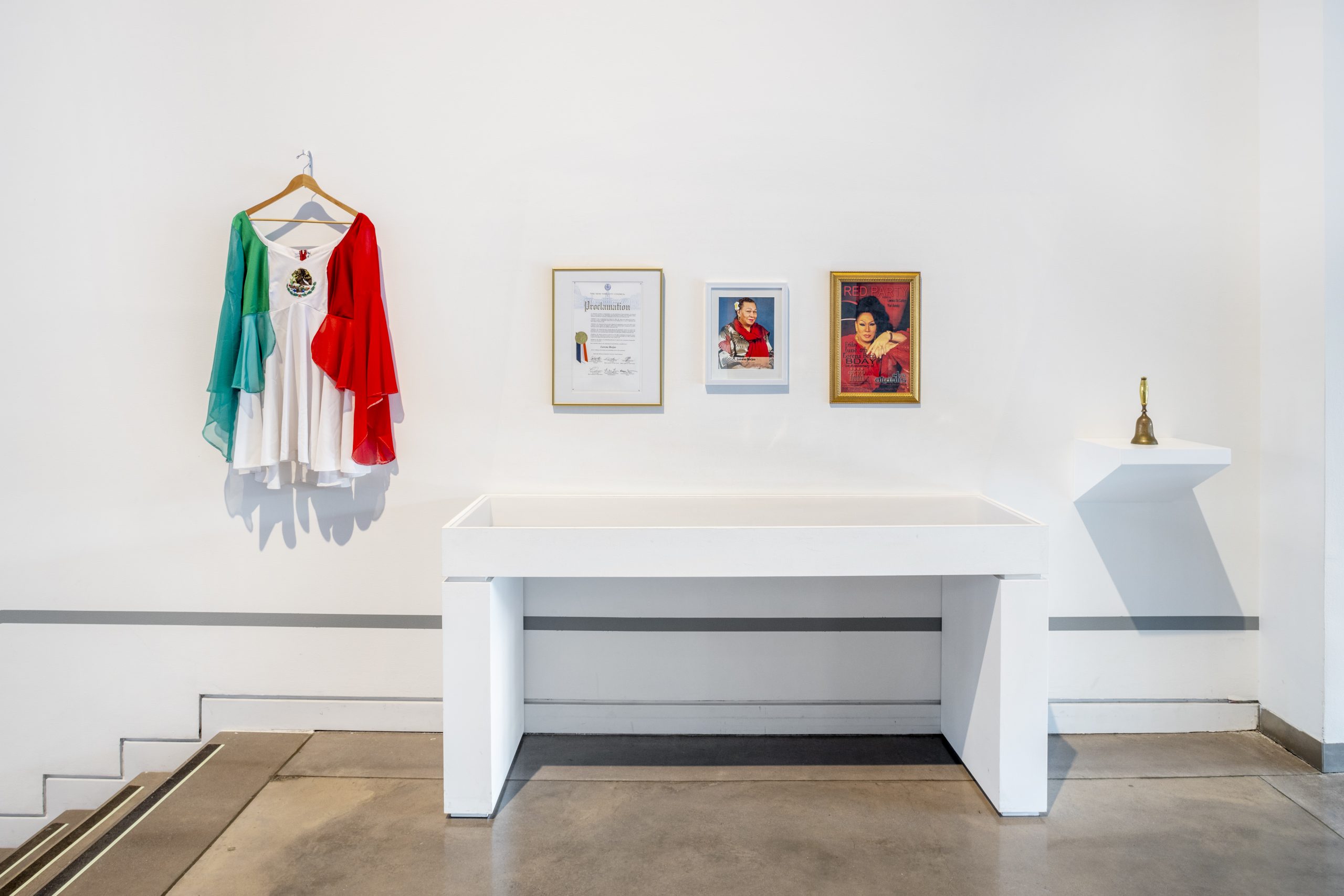 A white exhibition wall with a series of objects on display. Starting on the left is a Mexican flag fashioned into a dress hanging on a clothes hanger. Centered, is a framed certificate, a framed portrait of Lorena Borjas, and a framed magazine cover featuring Lorena Borjas. On the right is a gold handbell sitting on a white, exhibition shelf. In front of the wall is a long, rectangular display case.