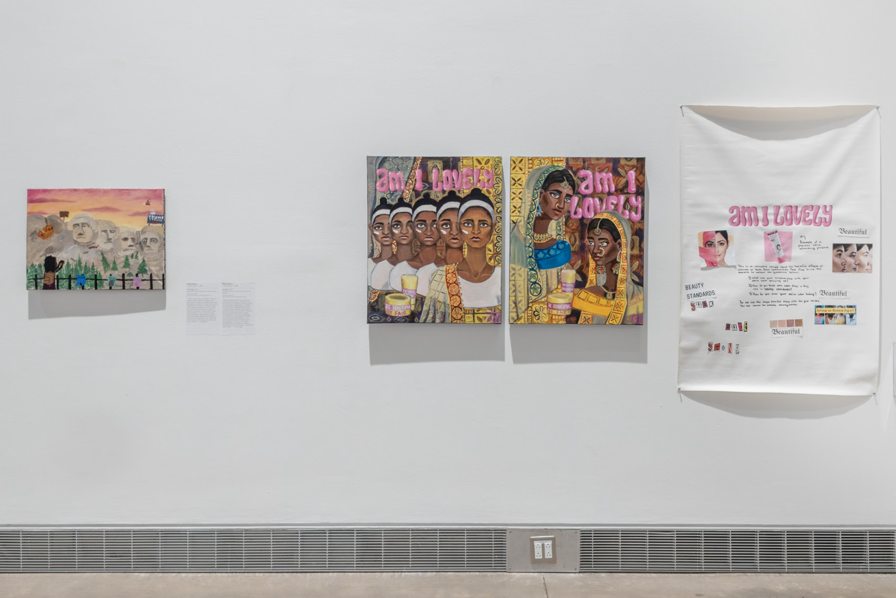 Paintings and a collaborative scrapbook hang on a white gallery wall. On the left, a small painting of Mount Rushmore at sunset, decorated with political protest signs, trees, and onlookers. On the left are two paintings with pink text: Am I Lovely? The left painting shows South Asian women of different brown complexions with white t-shirts and a white skincare product on their cheeks. The right painting shows two women in wrapped garments, gold facial jewelry, and head coverings. To the right, an interactive art piece asks the viewer to paste cut-out magazine clippings that explore colorism.