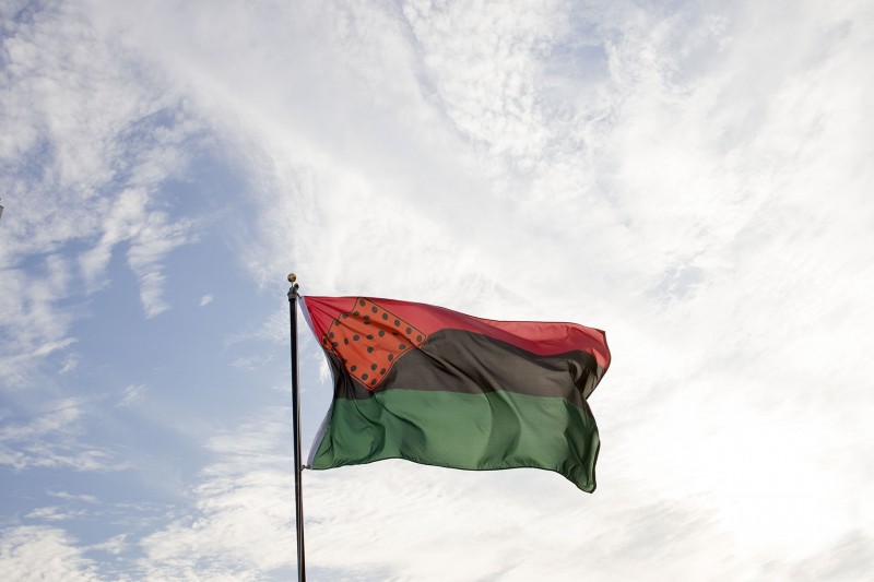 A wispy clouded blue sky in the daytime. Fluttering above it is a red, black, and green, horizontal striped flag on a black flagpole. In the top, left corner of the flag is a red diamond shape with black spots.