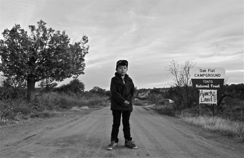 A black and white photograph of a Latinx boy standing in the middle of a dirt road. He is wearing dark clothing and a cap. On the right side of the road is a sign that reads “Oak Flat Campground, Tonto National Forest”. Taped on the sign is another sign that reads “Apache Land!!!” in spray paint.