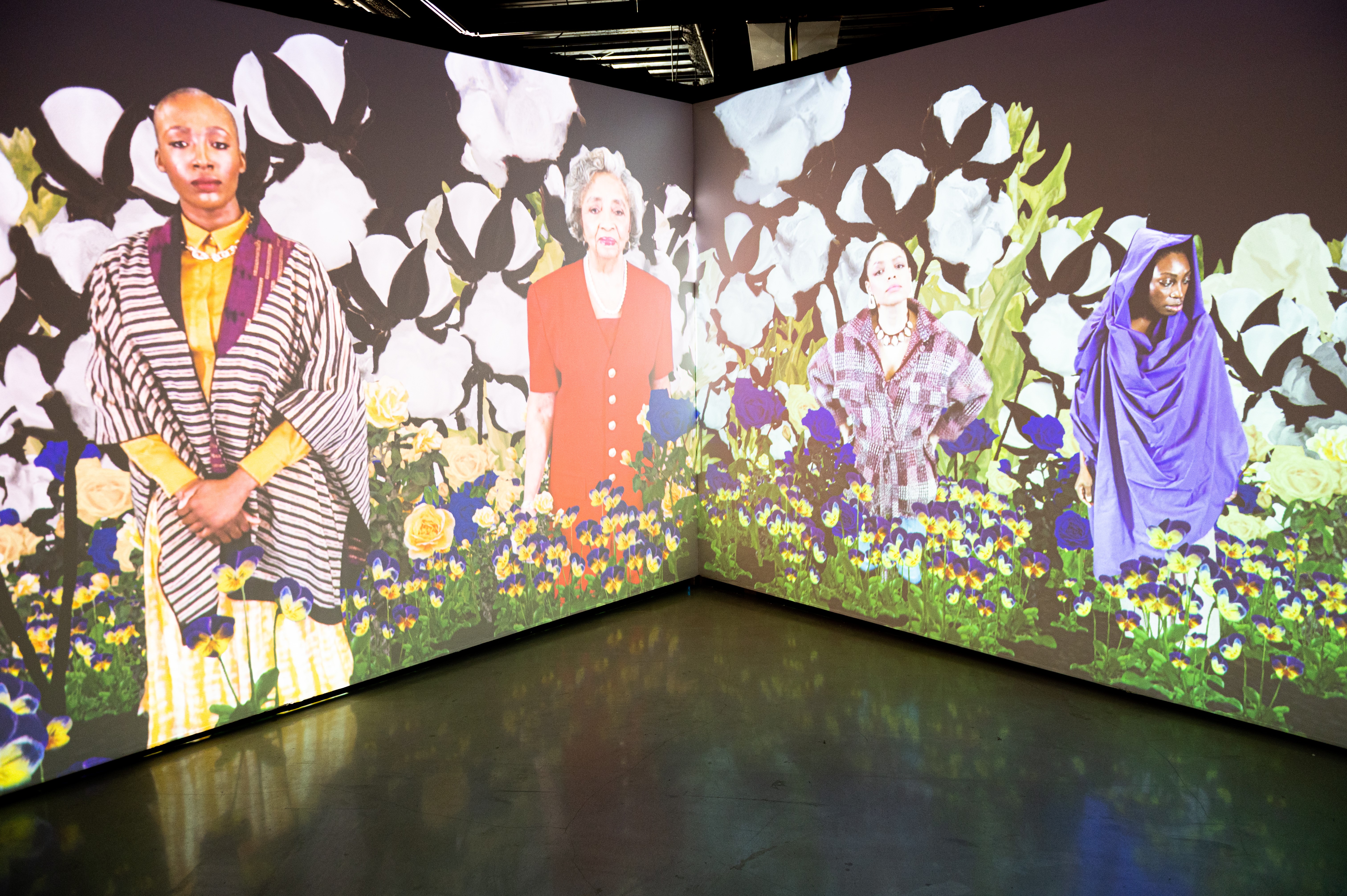 Video installation along two gallery walls depicts 4 women of color with various skin tones, age, and clothing surrounded by digital flowers & plants.