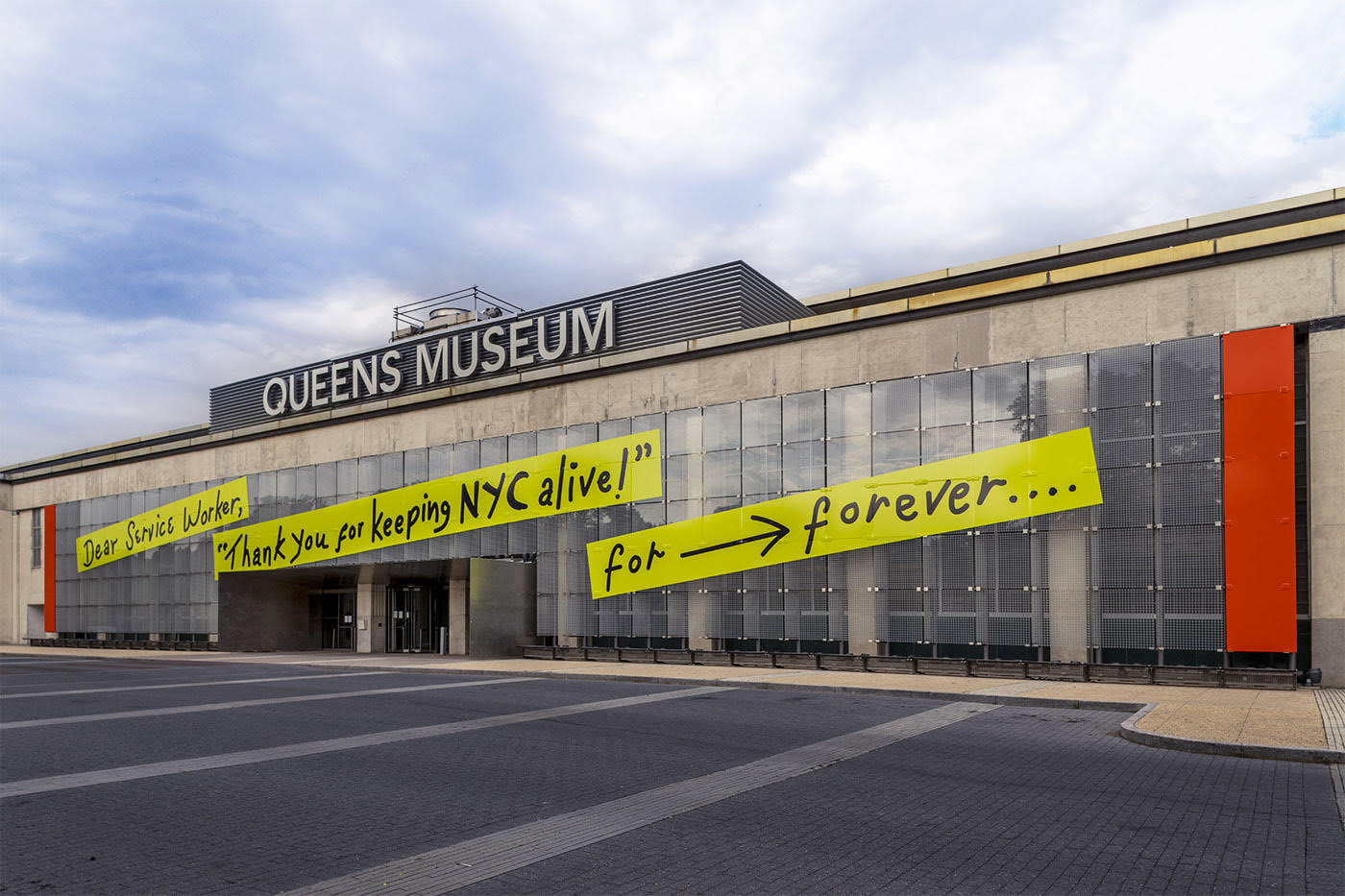 On the Facade of the Museum is a three part banner in yellow with the phrase written in black ink, Dear Service Worker, Thank you for keeping NYC alive! For forever… There is a forward arrow between the words for and forever. The top of the building says Queens Museum. All of this is set below clouds and blue skies.