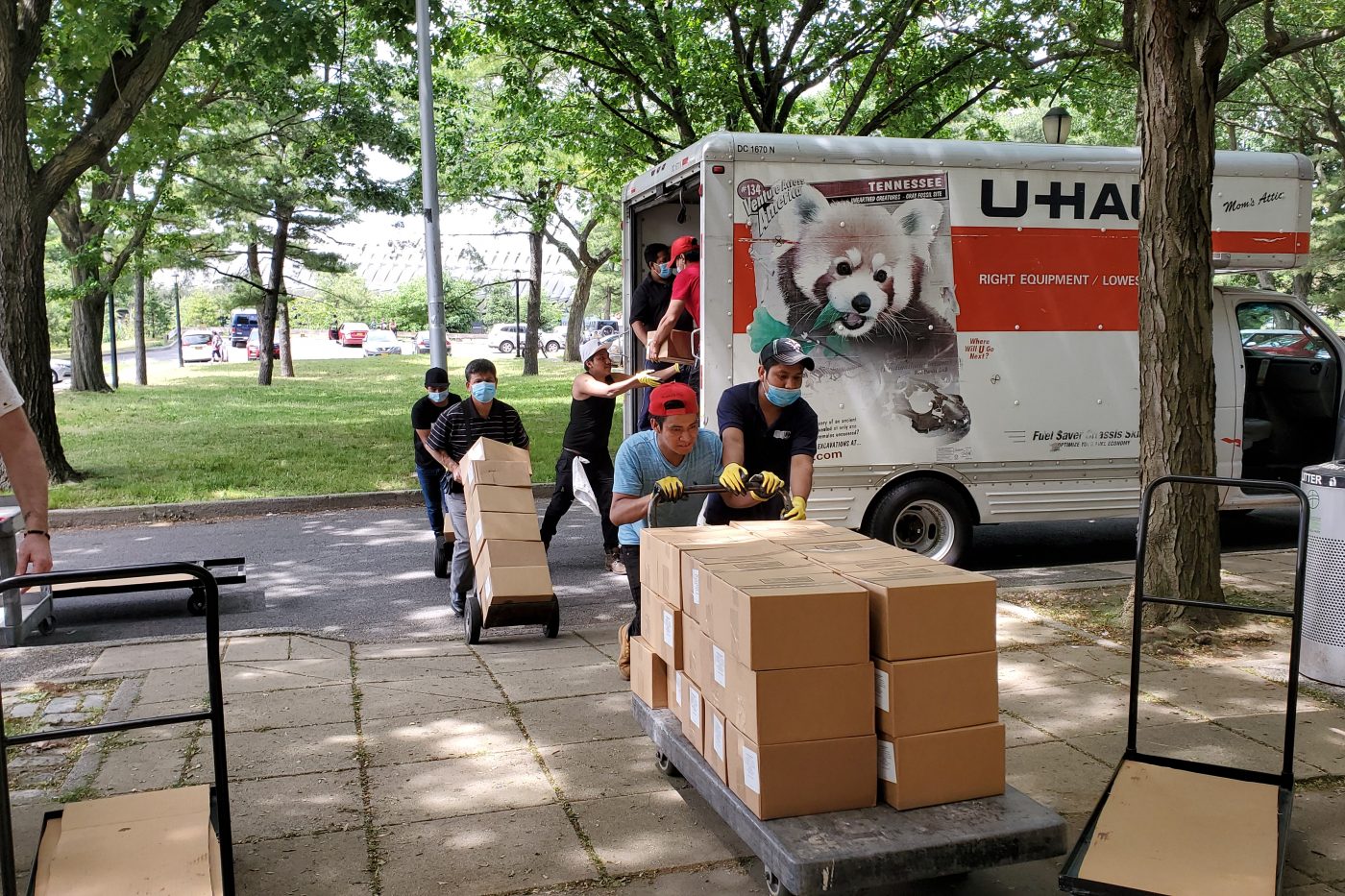 Seven Cultural Food Pantry volunteers are moving cardboard boxes from a parked U-haul truck to the Queens Museum's North entrance.