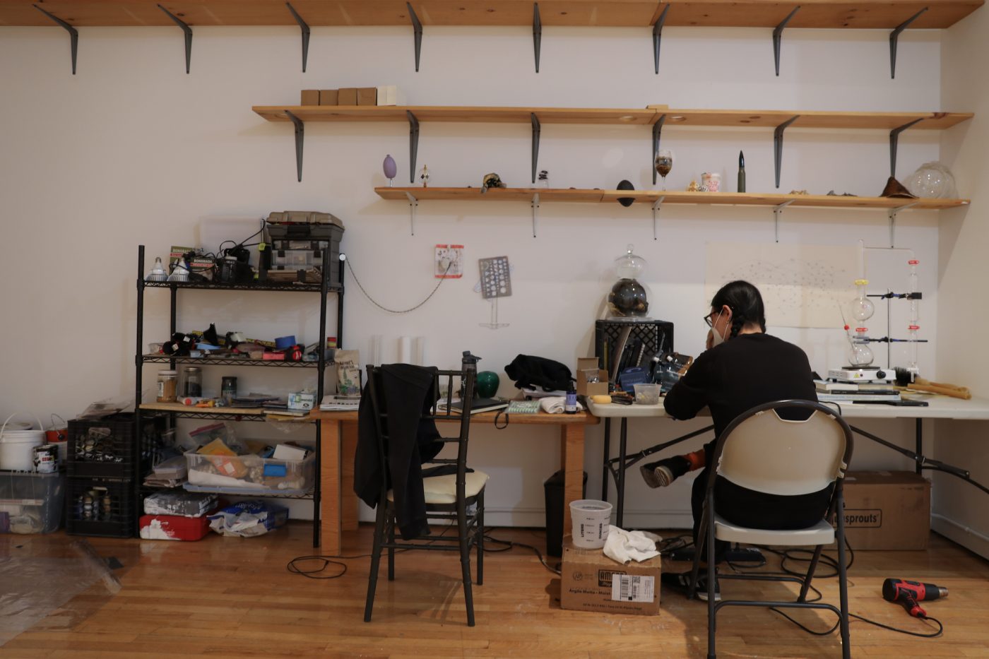 Artist Mo Kong is seated at their studio desk with supplies surrounding them and on shelves, including vessels resembling laboratory equipment.