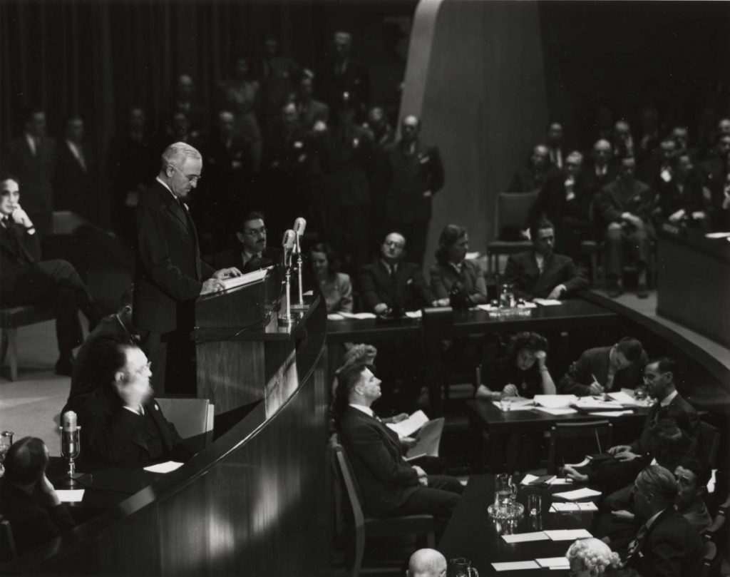 A black and white photo of a crowded room, filled with adults dressed in suits. Harry S. Truman, a mature, fair skinned man, is at a podium in the top left corner, addressing the crowd. Some of the adults are standing, some are seated at tables and taking notes, and some are seated in rows of chairs.