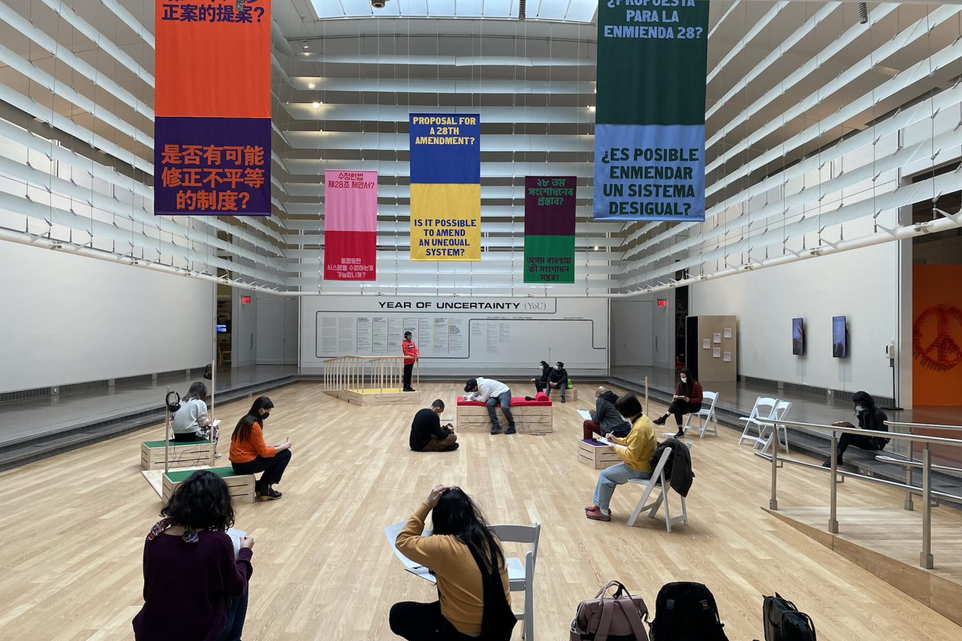Image of Alex Strada and Tali Keren's installation Proposal for a 28th Amendment? Is it Possible to Amend an Unequal System? presented in the Queens Museum's atrium. Five colorful banners are hanging from the ceiling, with the exhibition's title in different languages. Various visitors are engaging with the installation's different elements while sitting and standing.