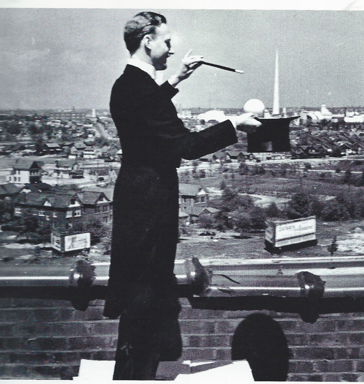 A black and white photograph of a white male, magician. He is standing on a platform and dressed in a tailcoat. In one hand he has a magic stick pointing towards his other hand, holding a top hat. In the background is Queens and the pavilions for the New York Fairs.