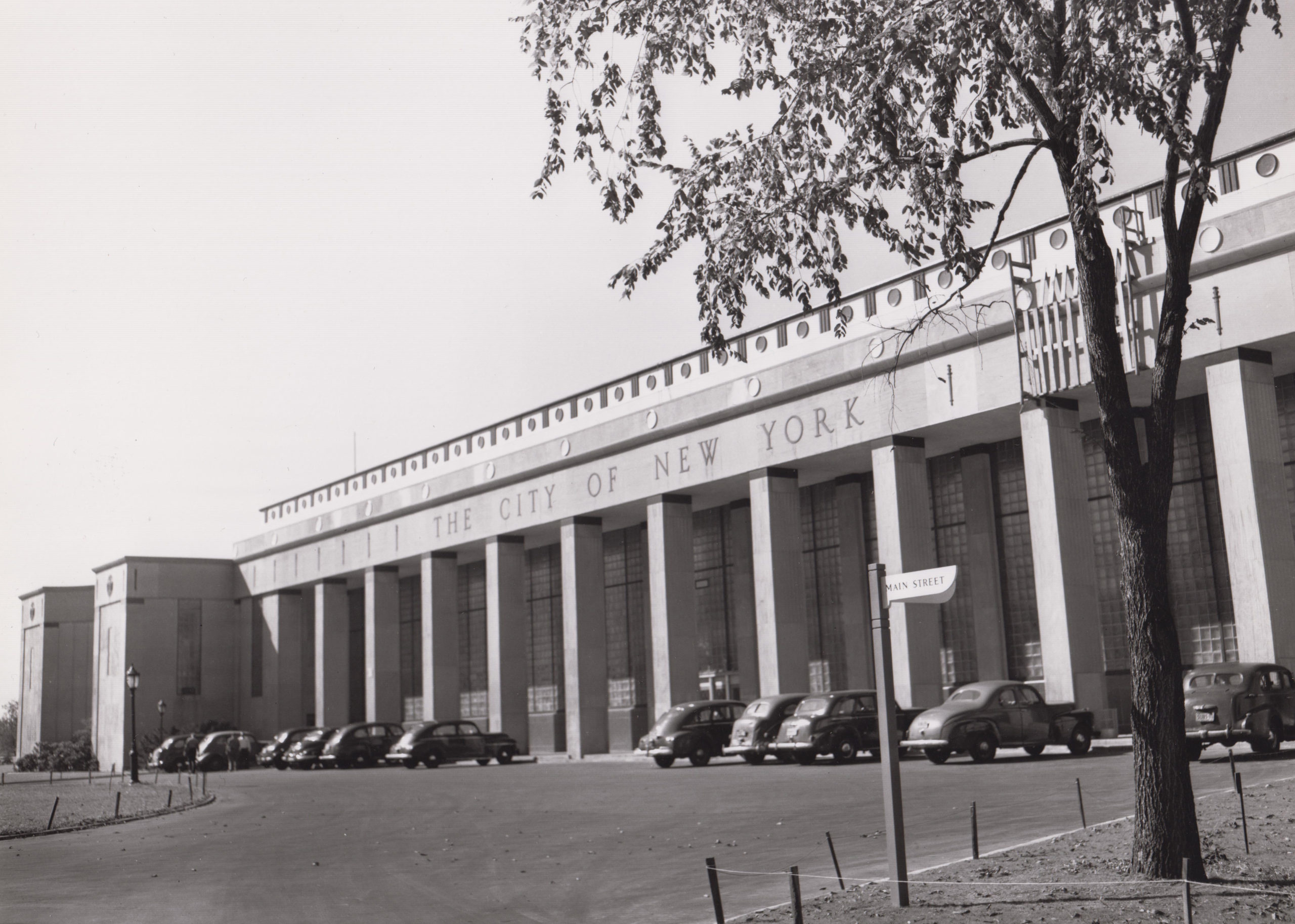 A black and white photo of the New York City Building. It has a long rectangular shape with a row of square columns. At the top of the building in uppercase font reads “THE CITY OF NEW YORK”. In the parking lot, in front of the building, are nine cars parked in a row and a street sign, to the right, that reads “MAIN STREET '' in uppercase font.