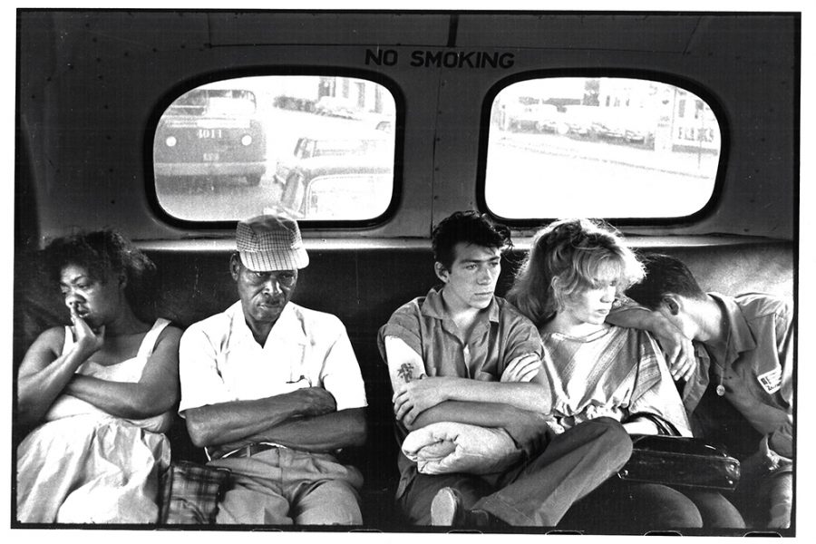 A black and white photograph of five people sitting in the back row of a bus. The people are of different racial backgrounds and genders. Their arms are crossed and they are sinking into the back seat. The fatigue of public transport is written on their face. Behind them are two round windows framing road they are passing over and a sign the reads “No Smoking” in all caps.