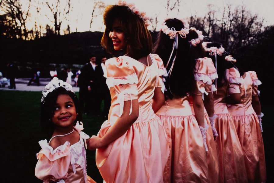 A color photograph a bridal party on a park lawn. Descending from the center is a row of bridesmaids dressed in peach colored ,90s era dresses. The dresses have a low ruffled skirt and puffy shoulders with white, lace trim. Each bridesmaid is wearing matching white, lace gloves and peach, floral crowns, with the exception of the bridesmaid closest to the camera. She and a flower girl are both peering over their shoulders smiling.