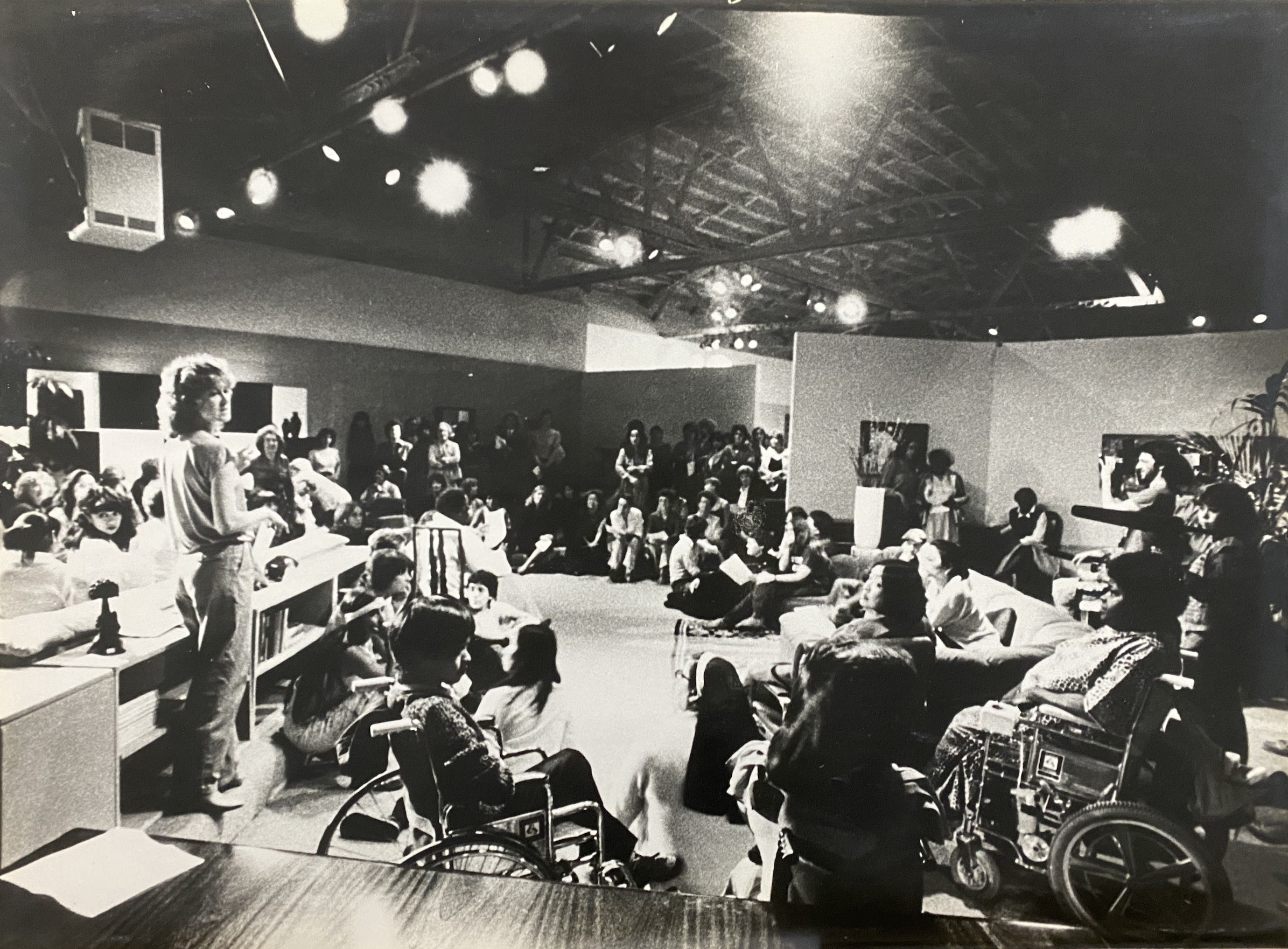 In a black and white image set in the 1970’s, Suzanne Lacy stands to the left of the frame overlooking a room full of people. At the forefront are a group of people in wheelchairs, along the sides people are sitting on a couch. In the background people are standing along the room while others are sitting on the floor creating an oblong empty space in the center.