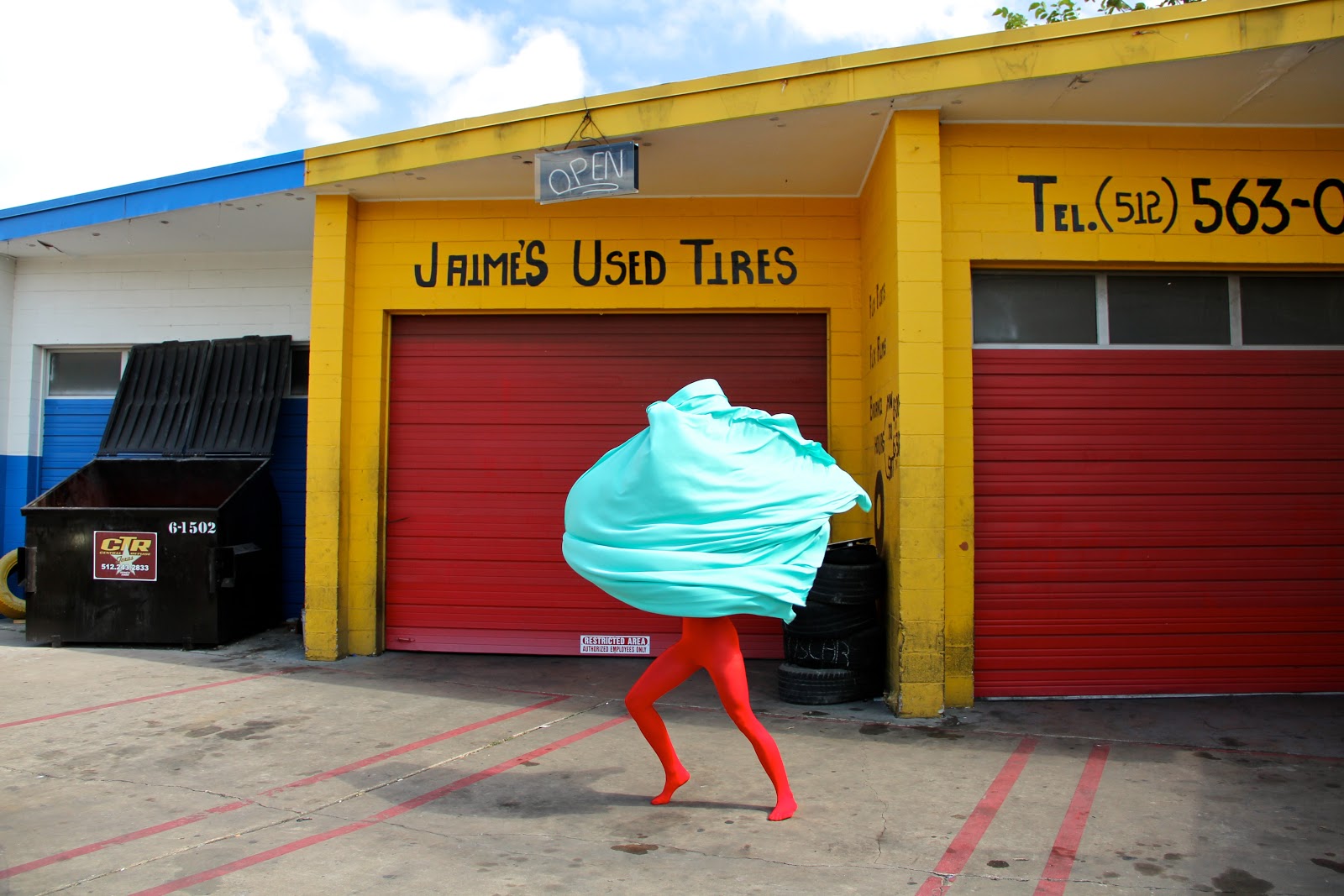 A figure in a red bodysuit and large bundle of icy blue fabric covering the top half of their body walking across a driveway. In the background is a yellow building with red garage doors and a sign that reads “Jaime’s Used Tires”.