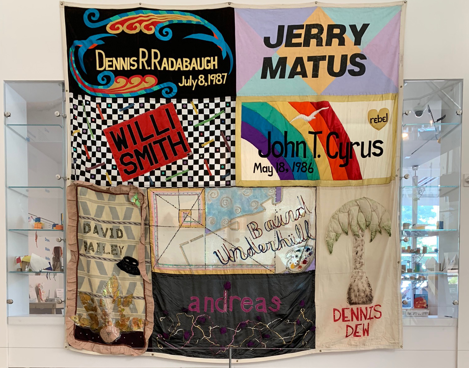 A large quilt hangs from ceiling of an exhibition wall. The square quilt is made up of eight rectangular patches. Each patch is made up with different patterns, colors, and motifs and has the name of a community member lost to AIDs in large font.