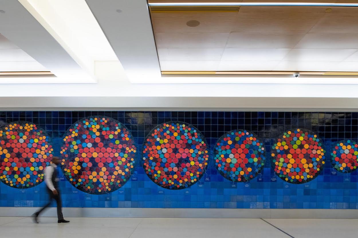 A video thumbnail with a centered, black “press play” symbol in a green rounded off square. The thumbnail is of a blue-gradient tiled art installation, covered with six large multi-color circles. In front of the art installation a dark skinned man walks by.