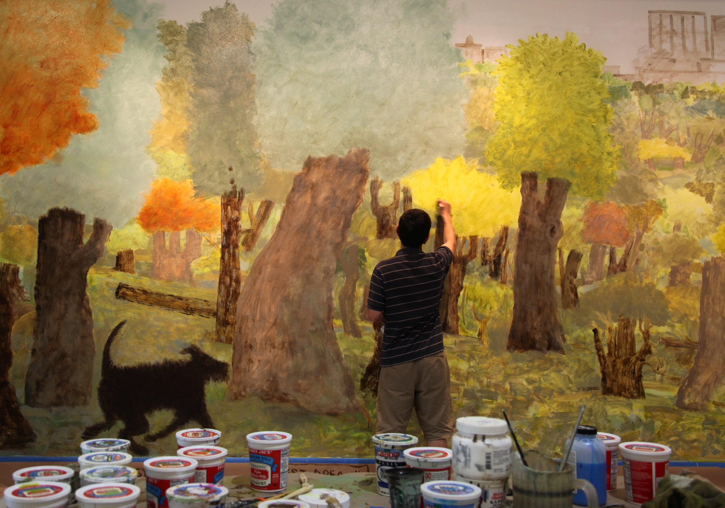 In the foreground is a table surface with recycled, yogurt containers filled with paint, paint brushes, and a pitcher of water. In the background, the artist David Kearns, has his back turned and is painting a mural made up of trees transitioning into fall colors. The mural features a black-haired dog walking amongst the trees and a city scape in the background. The paint style is blotchy and fuzzy with details not completely made out.