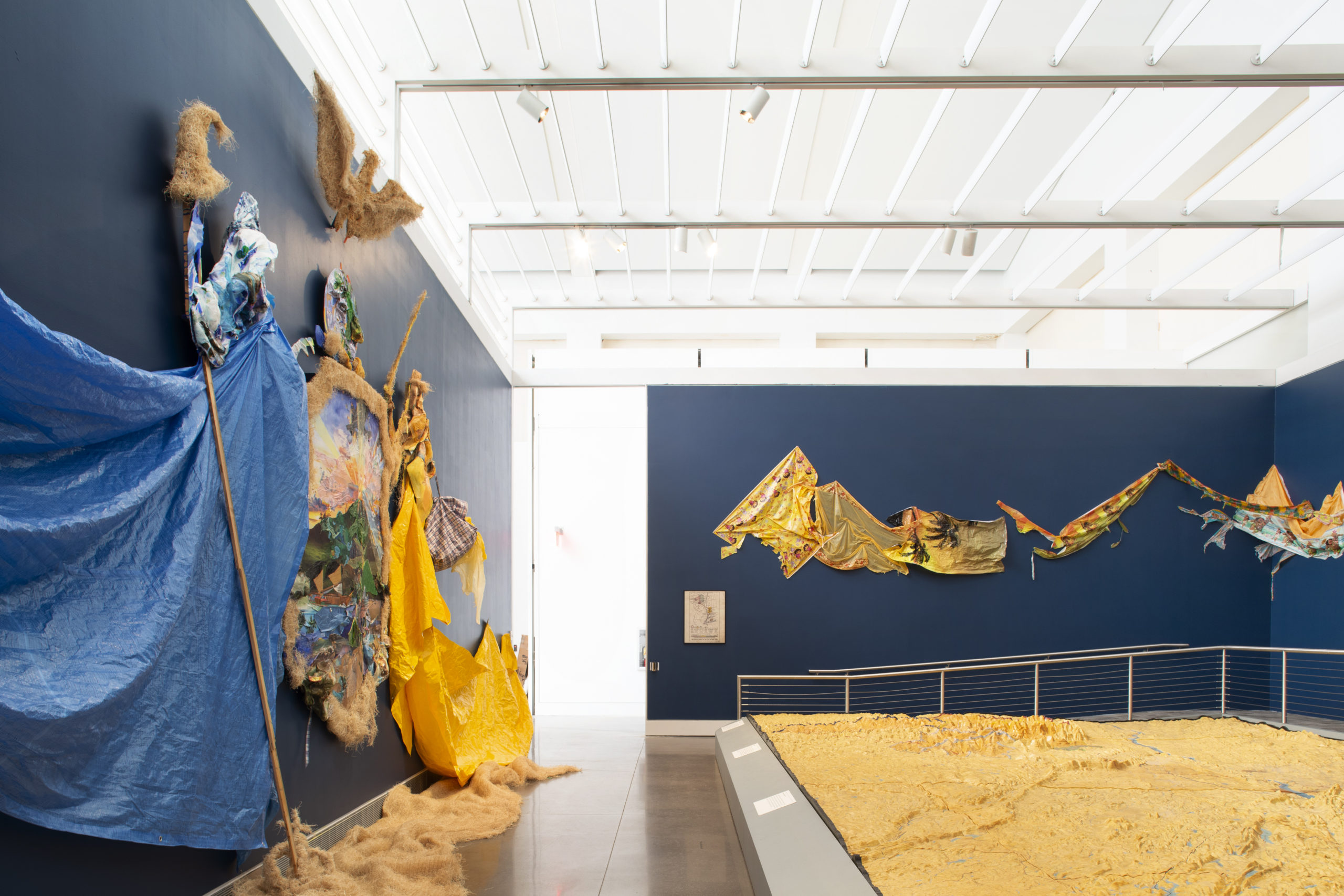 A exhibition space with a relief map of New York City’s water system installed on the floor. Surrounding the relief map is navy blue walls with a mixed-media collage and painting installation. The installation has an organic shape and is made of mostly blue, brown and yellow tones.