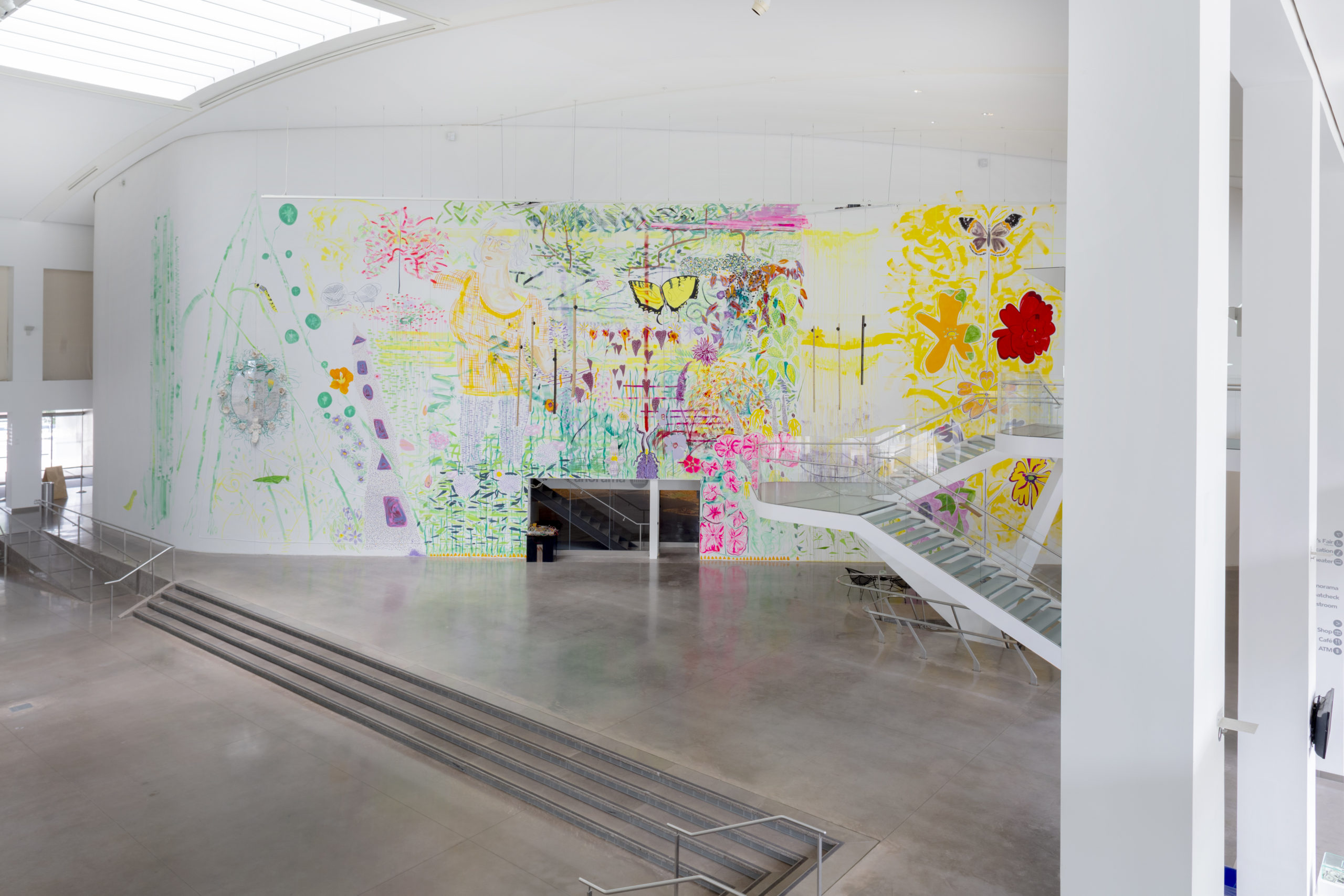 A two-story mural, of abstracted garden patterns and imagery in an empty exhibition space. Descending from the top right corner is a twisting flight of stairs. The first two-thirds of the mural, starting from the left, is mostly green and depicts a person standing in a field. As you move towards the right you see flowers in warm colors and a yellow butterfly. Then the green abruptly stops and a yellow scene begins with more flowers in different colors and a brown butterfly.