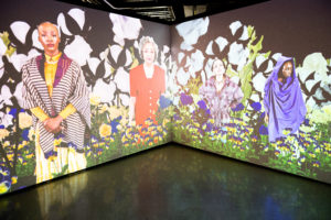 A projection covering two walls, coming together to create a corner. On the screens are four Black women, standing in a garden with purple and yellow flowers at their feet. Behind and around them are white flower petals. One woman wears a striped jacket over a yellow dress. Besides her an older woman in a red suit, next to her, a young woman in a plaid shirt. The final woman wears purple fabric draped over her head and body.