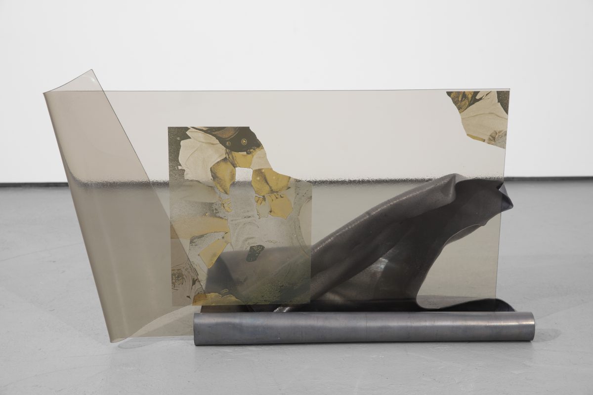A gray colored almost transparent rectangular piece of glass with a fold on the left side sits inside of a silver lead cylindrical folded base. The folds are soft . Inside the glass is an abstract shattered image in a rectangular shape with the top corner cut and placed at the far corner of the glass. The folds of the silver stand are visible through the glass.