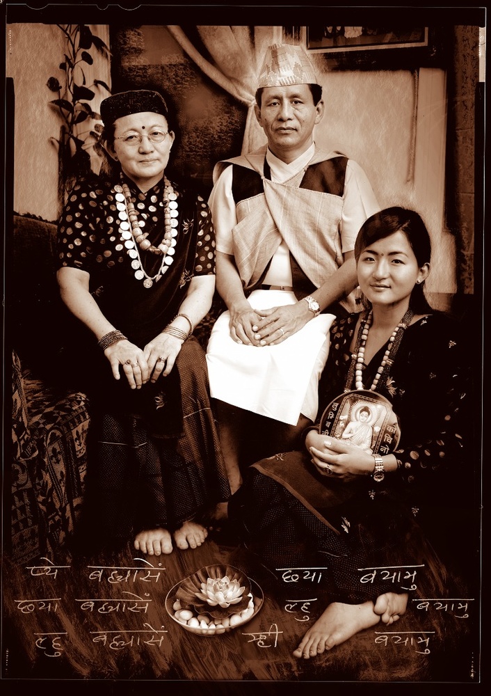 A black and white family portrait of a middle-aged man and woman, and a young woman. They are sitting in a living space and dressed in traditional Gurung clothing. The young woman is holding a round object with an illustration of a religious deity. At all three of their feet sits a bowl with pebbles and a lily-pad. Written on the photograph in white is Gurung script.
